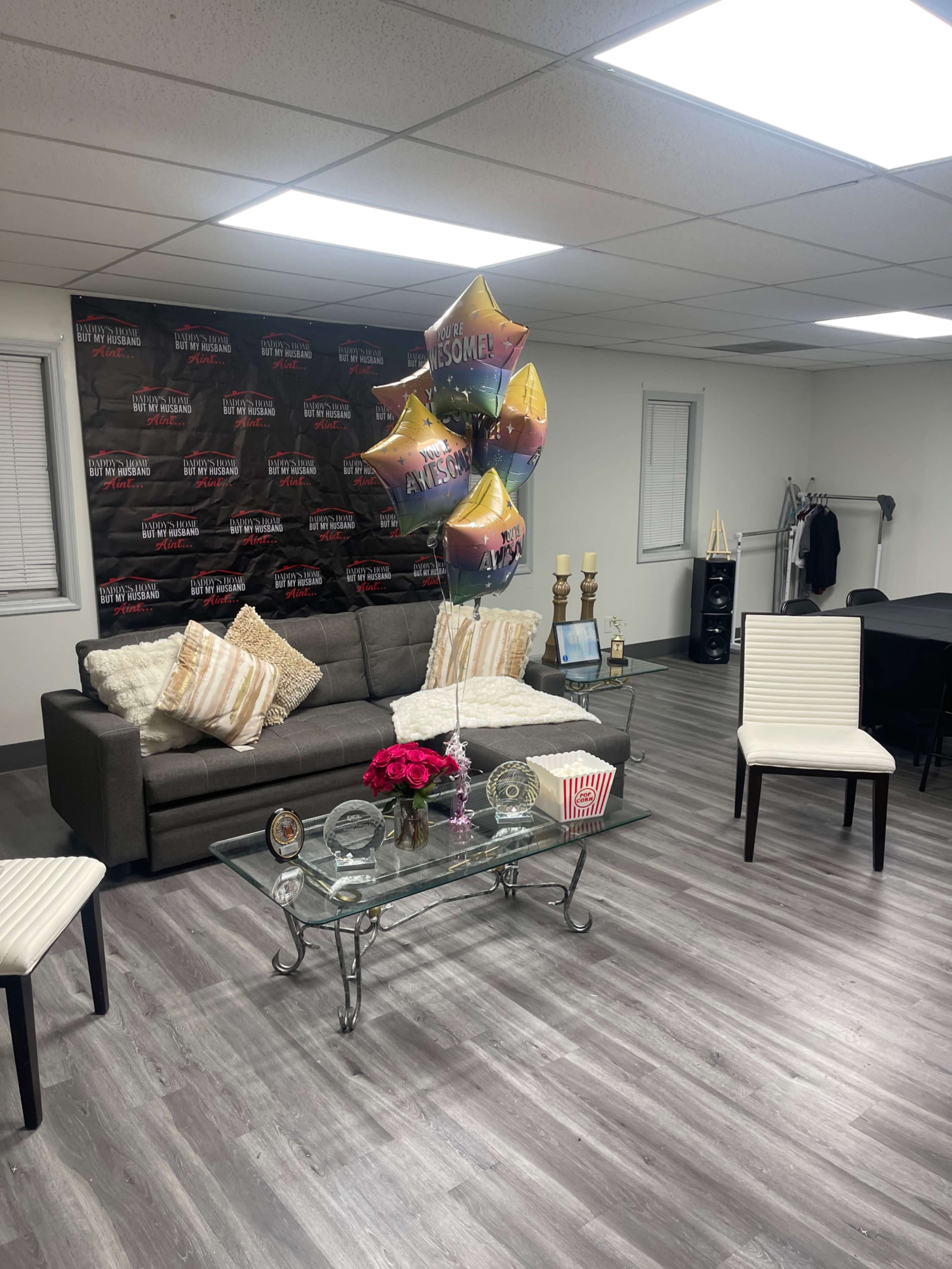 The image shows a cozy lounge area with a gray sofa, white chairs, a glass coffee table with a bouquet of flowers, and colorful balloons, set against a backdrop featuring a promotional display.