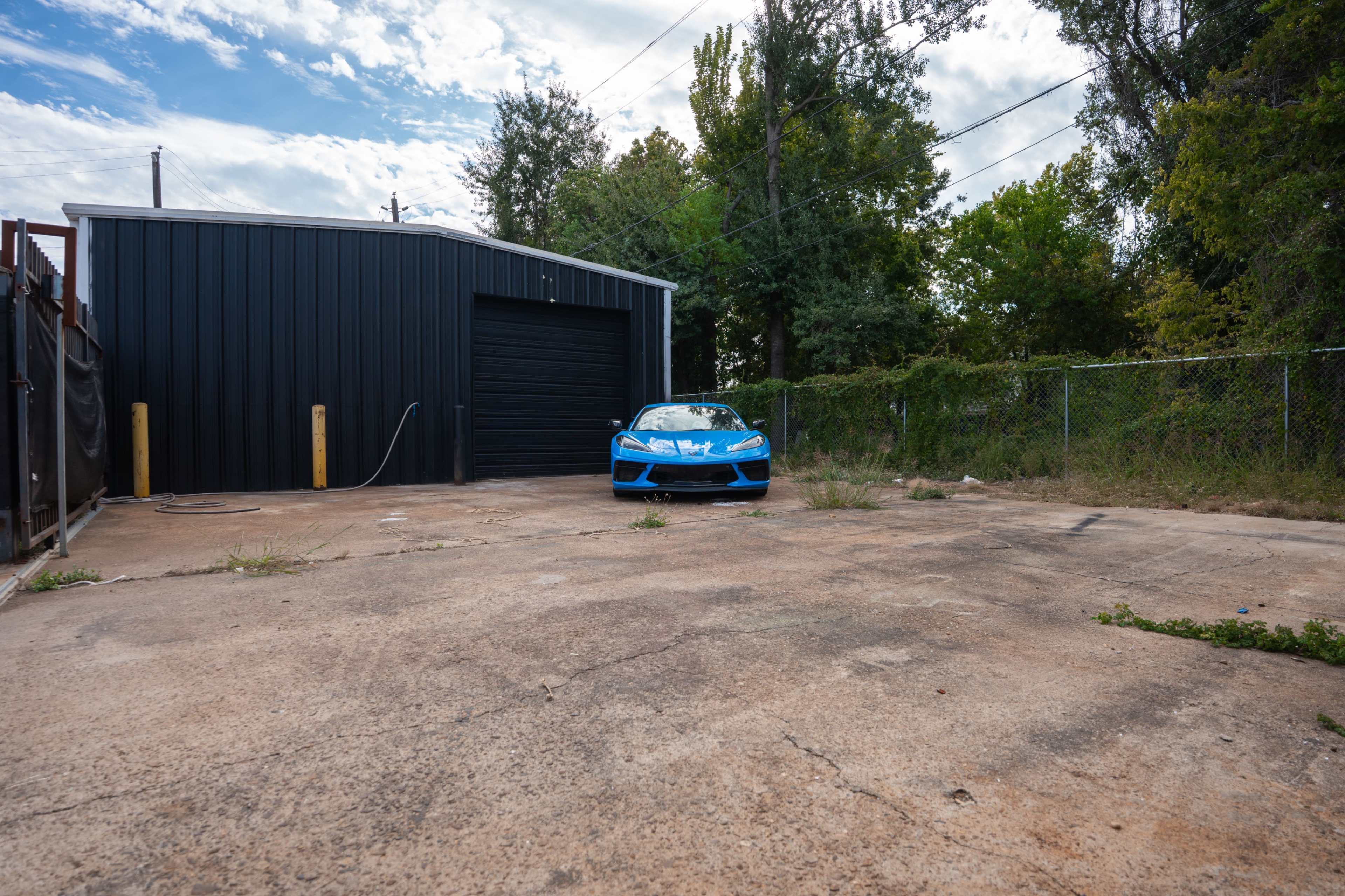 A bright blue sports car is parked on a concrete slab in front of a black metal building surrounded by overgrown grass and trees.