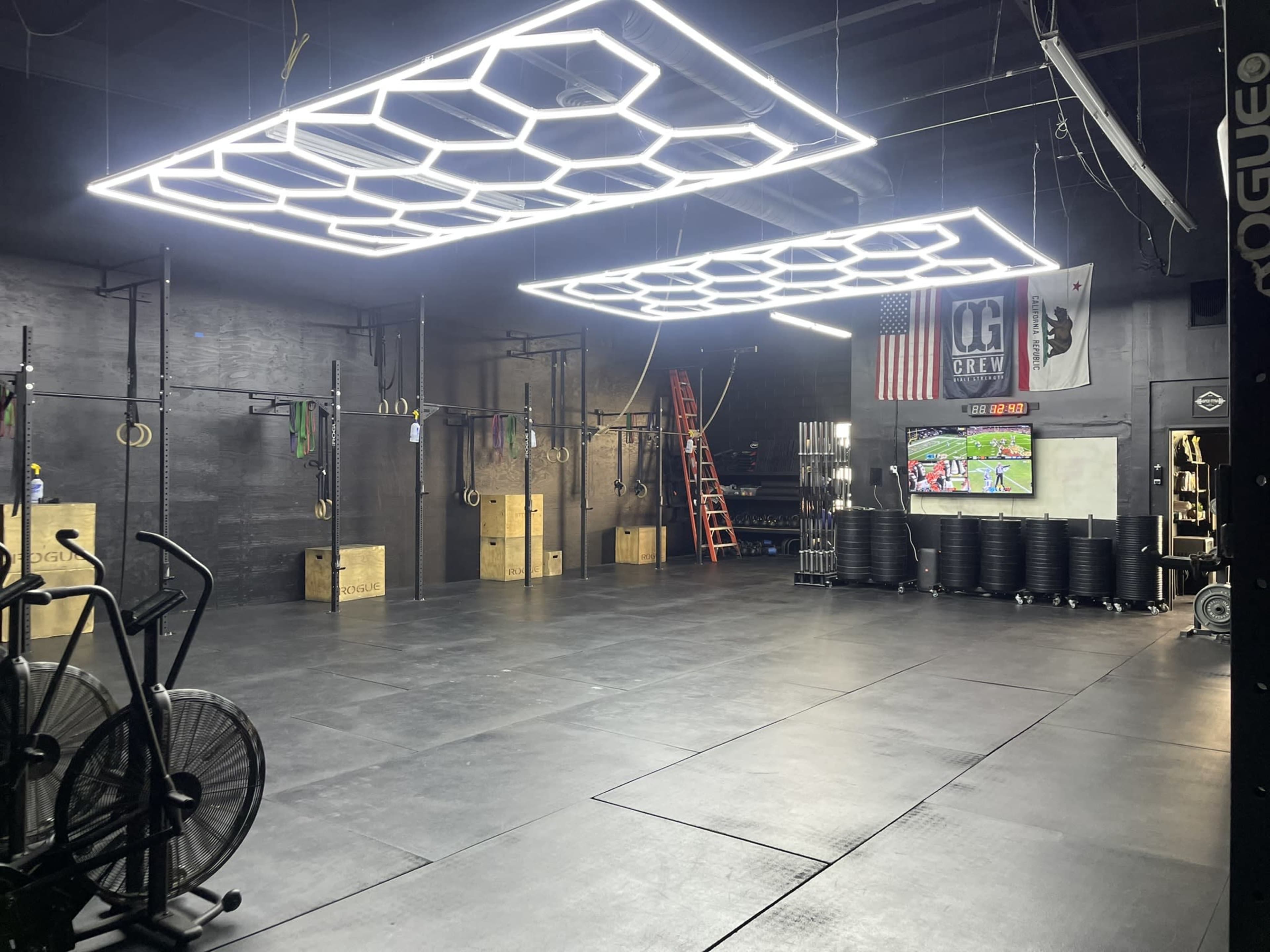 A gym interior featuring exercise equipment, high ceilings with geometric light fixtures, and storage boxes, with an American flag and workout-related banners on the walls.