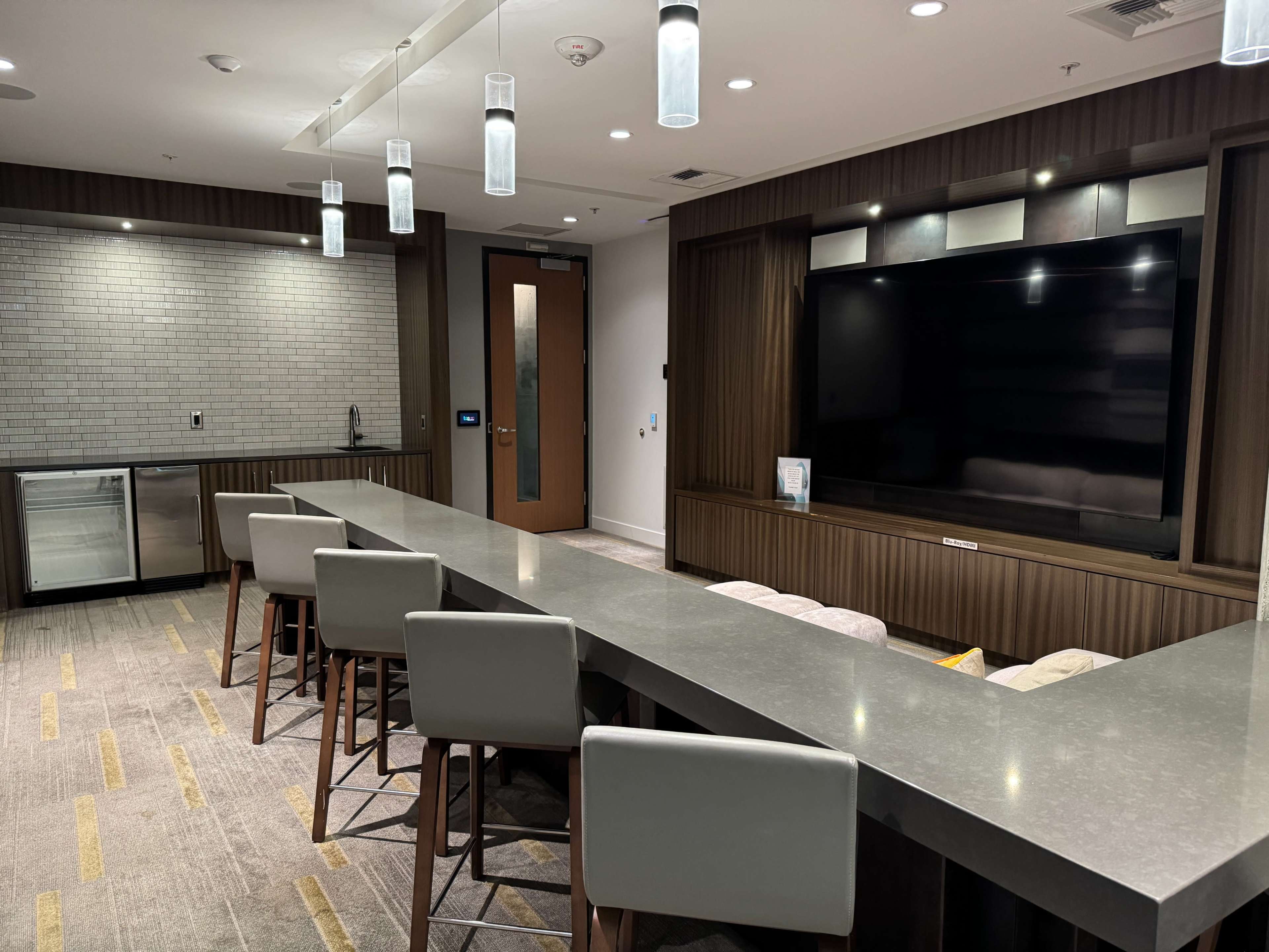 The image shows a modern meeting room featuring a long gray table with bar stools, a wall-mounted television, and a kitchenette area in the background.