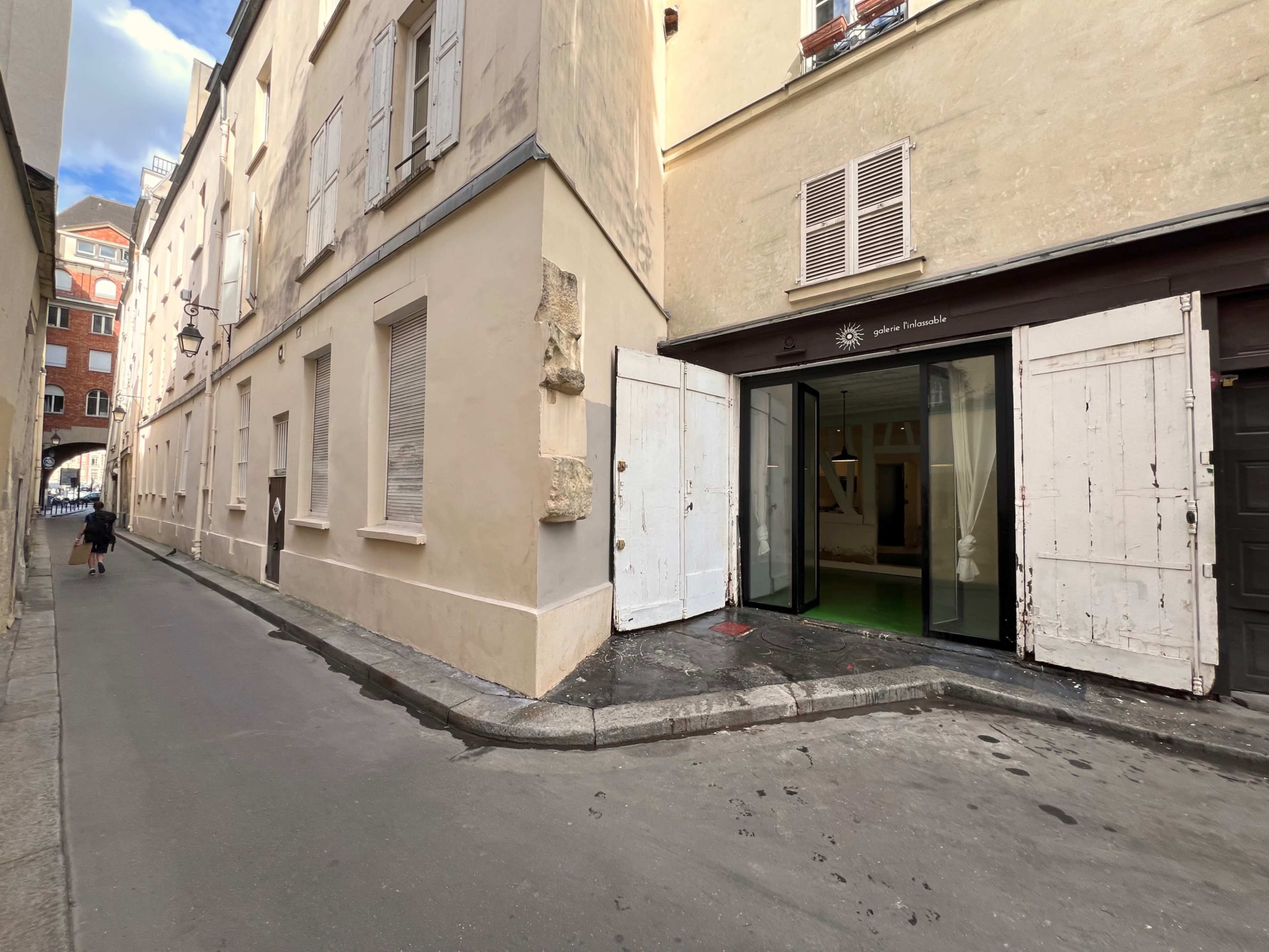 A narrow street with beige buildings and an open entrance featuring large glass doors on the right.
