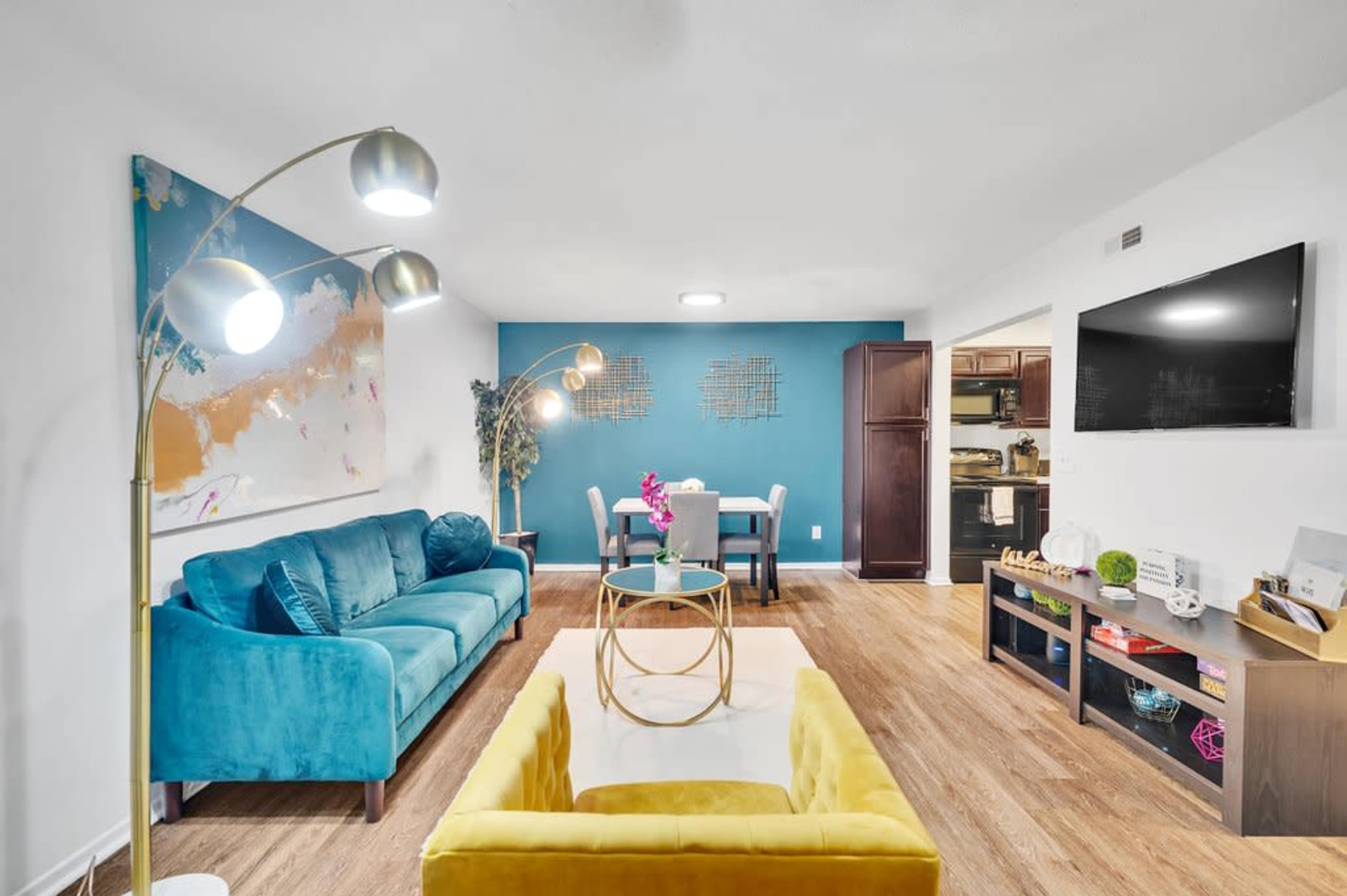 A modern living room featuring a teal sofa, a yellow armchair, a round coffee table, and a dining area with a light blue wall in the background.