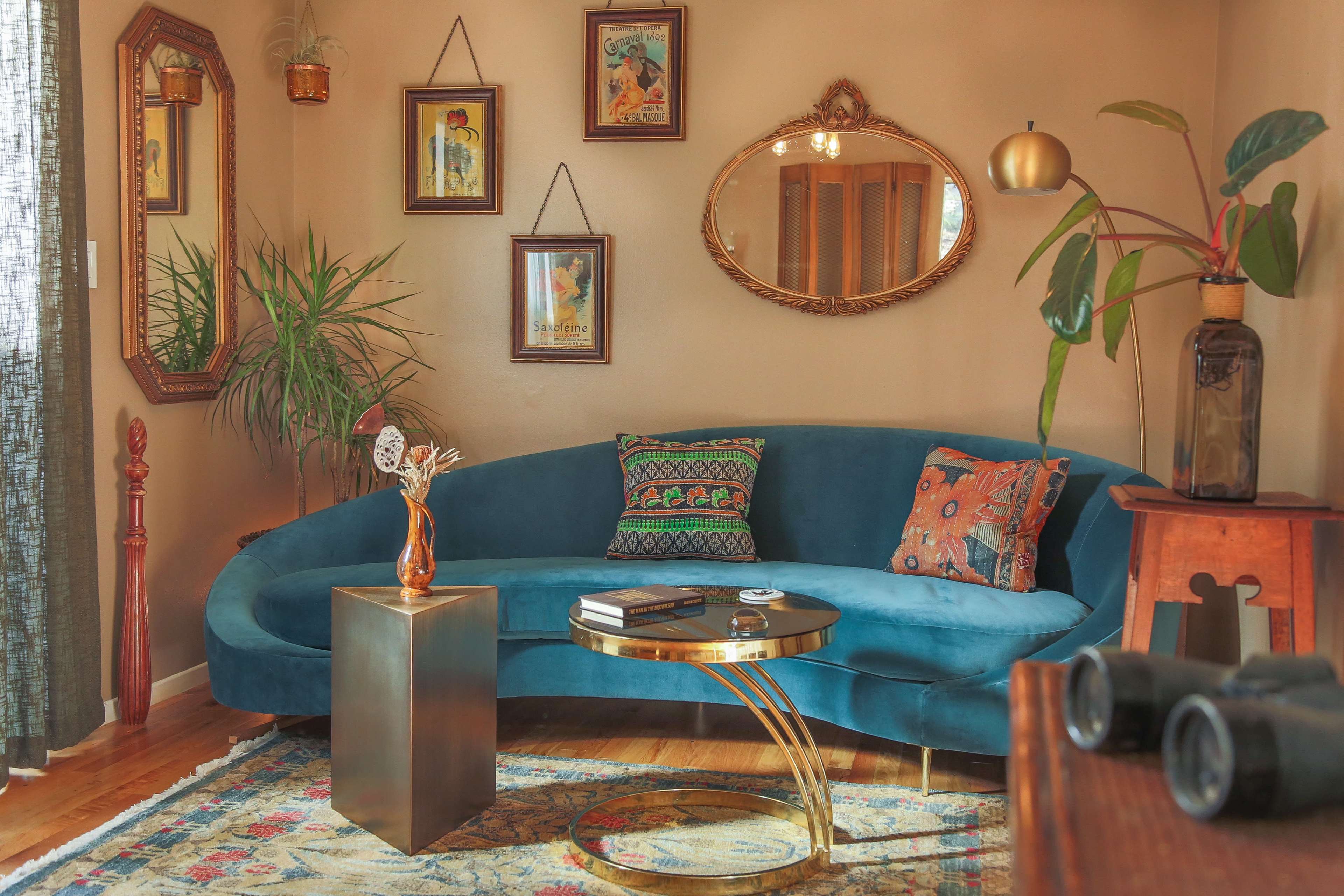 The image shows a cozy living room with a blue, curved sofa, a gold coffee table, and various decorative plants and framed art on the walls.
