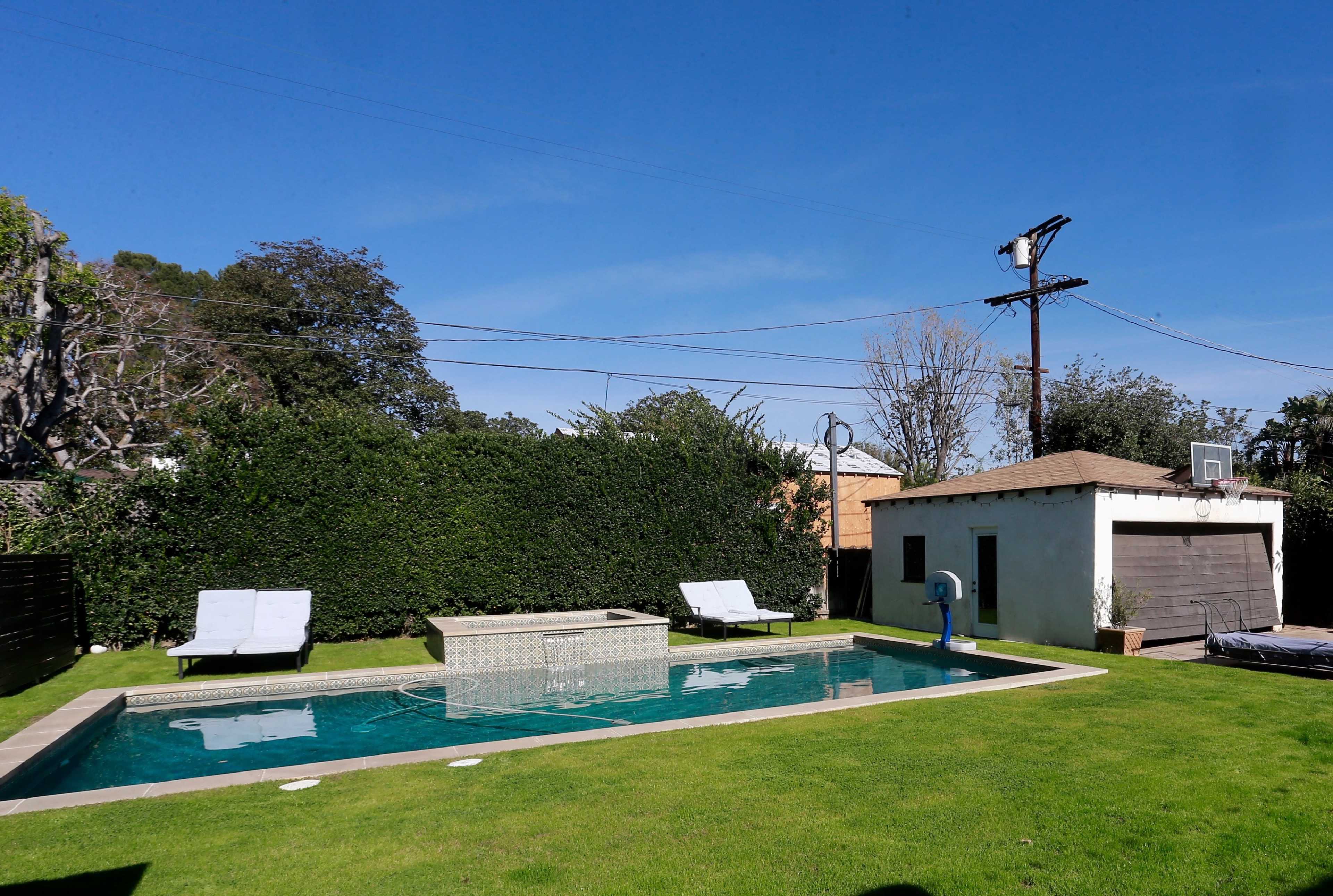 A backyard with a rectangular swimming pool, two lounge chairs, and a small shed surrounded by grass and trees under a clear blue sky.