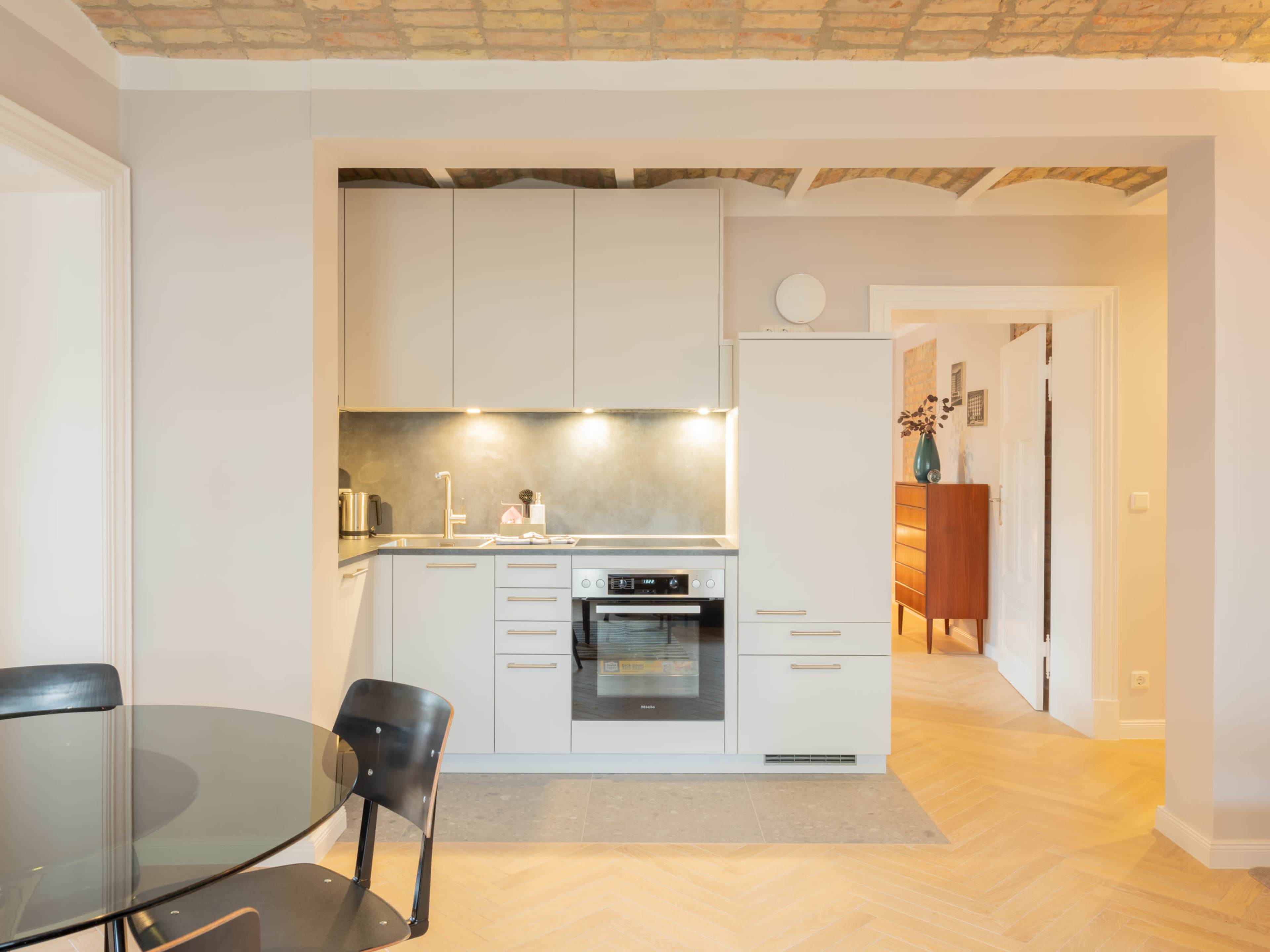 The image shows a modern kitchen adjacent to a dining area, featuring light-colored cabinetry, a stove, and a glass dining table with black chairs.