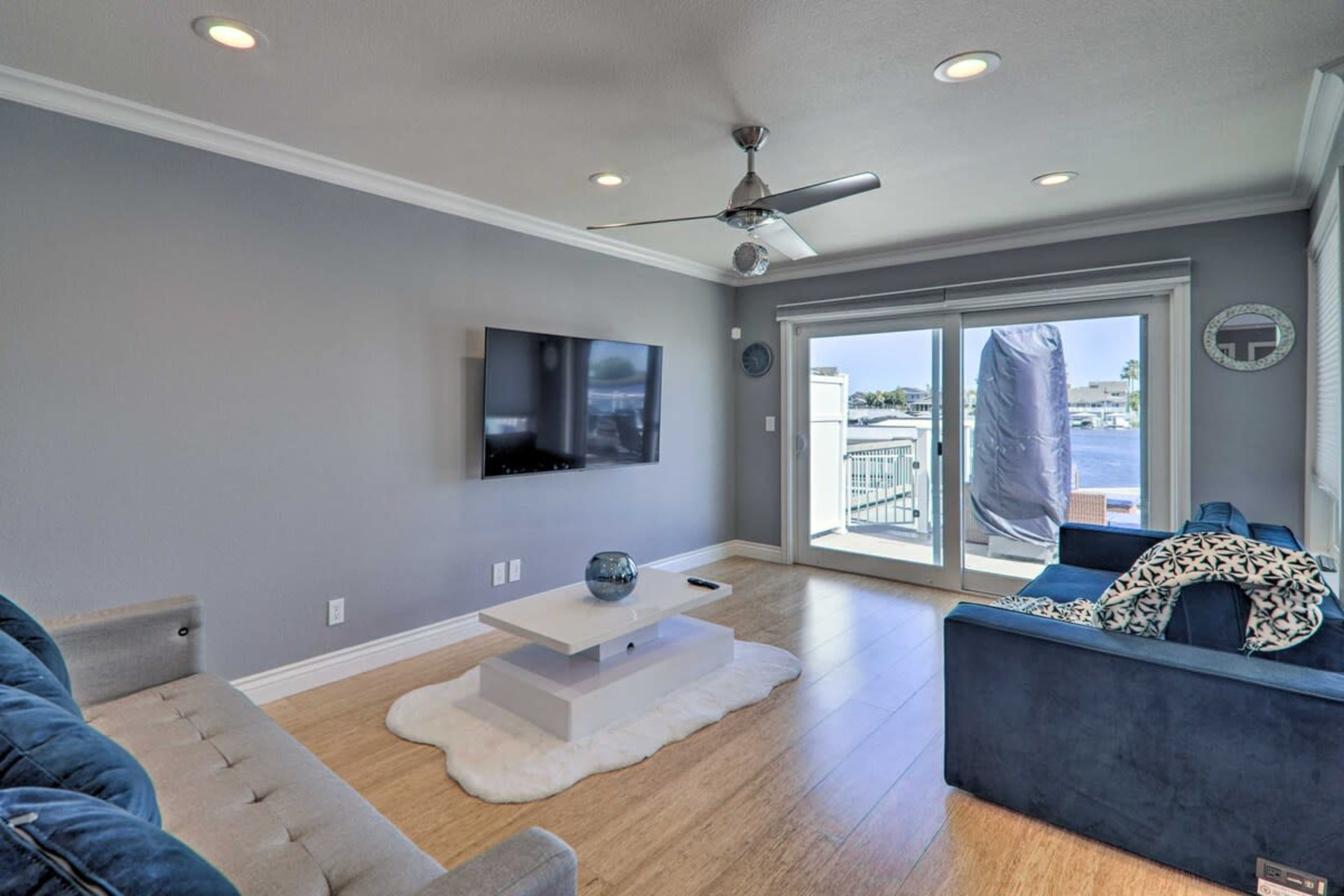 A modern living room features a flat-screen TV mounted on a light gray wall, a white coffee table on a rug, and large windows showcasing an outdoor view.