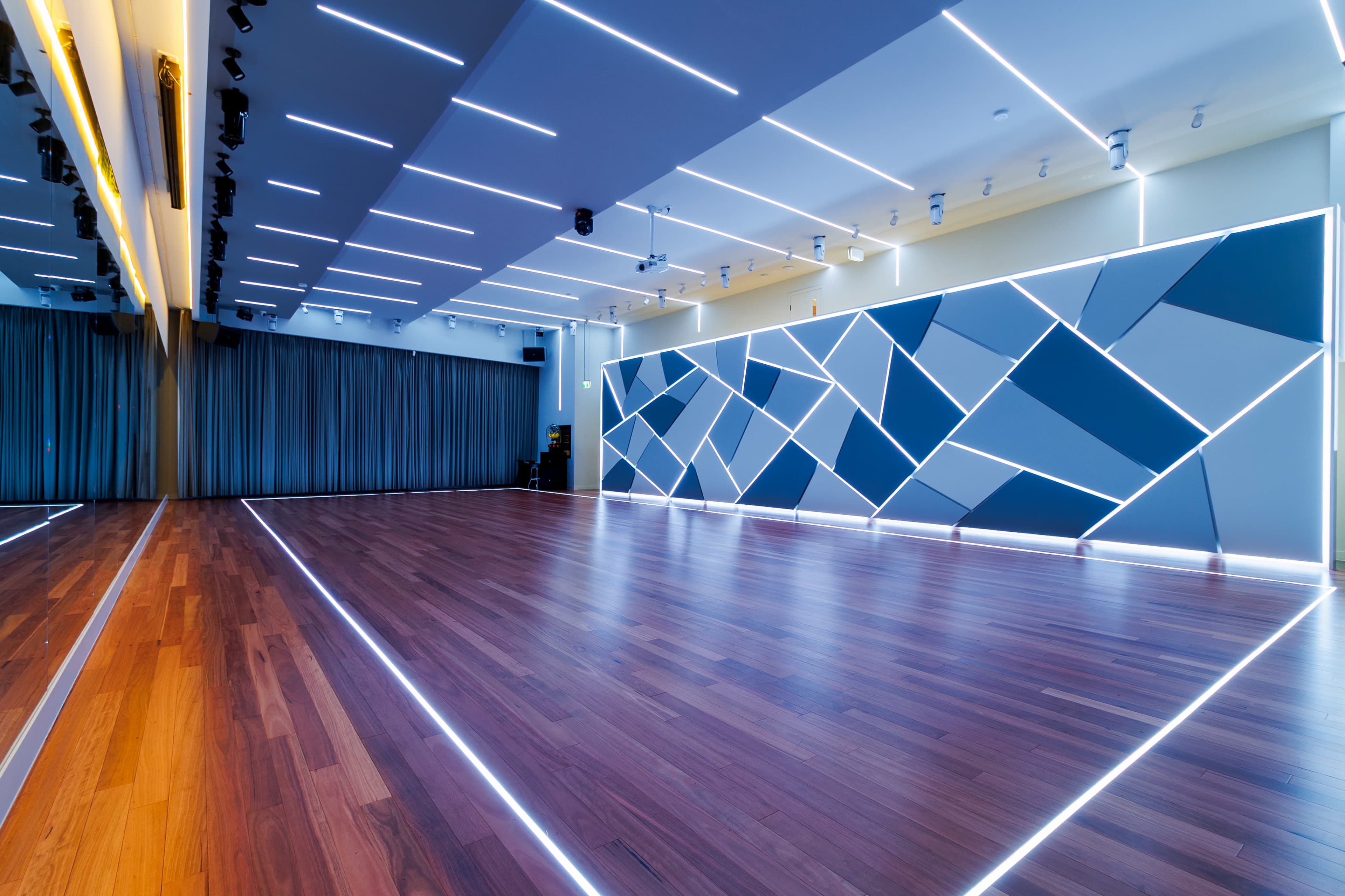 The image shows a modern dance studio with wooden flooring, illuminated by blue and white LED lights, featuring geometric wall designs.
