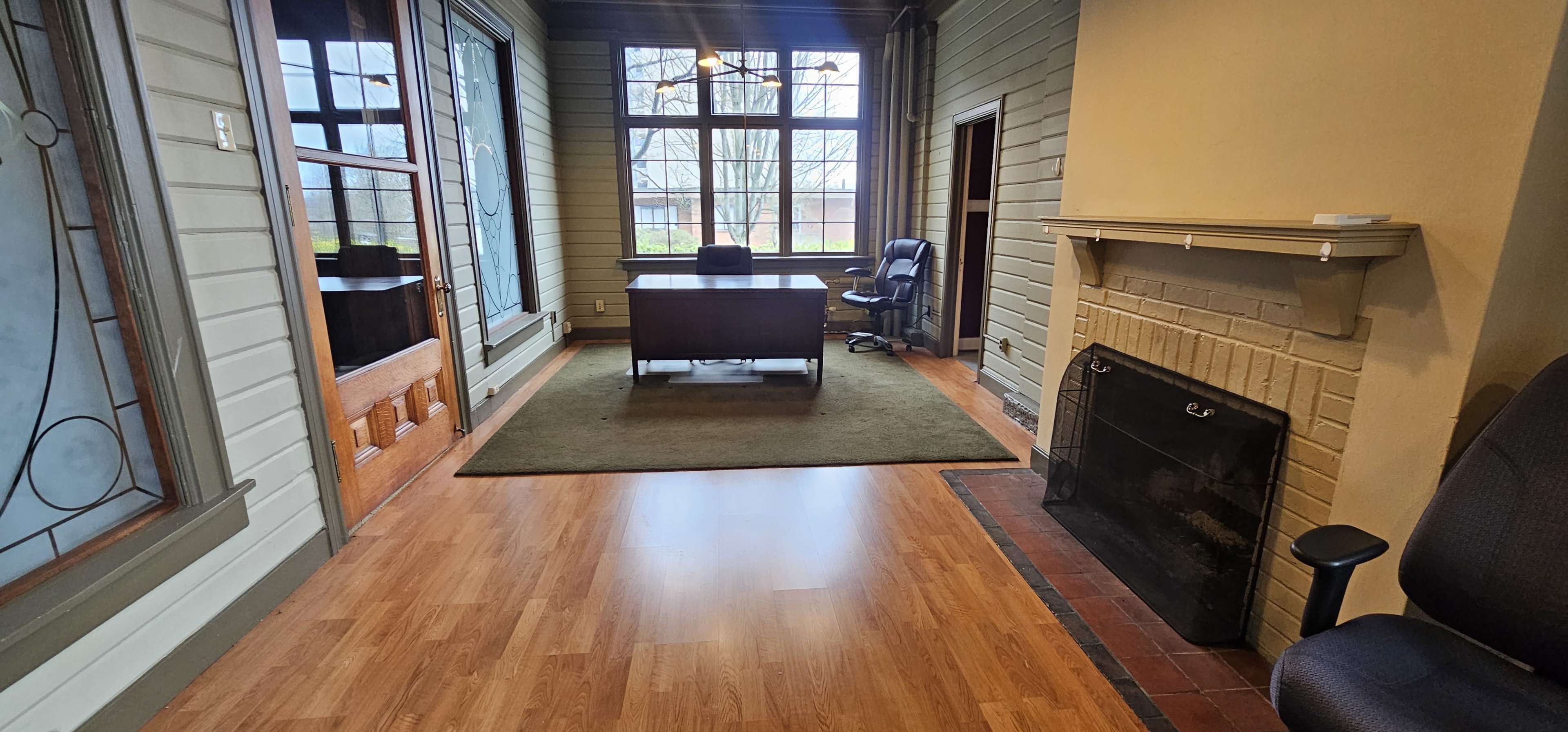 An office space featuring a wooden desk with a chair, a fireplace, and large windows allowing natural light to enter.