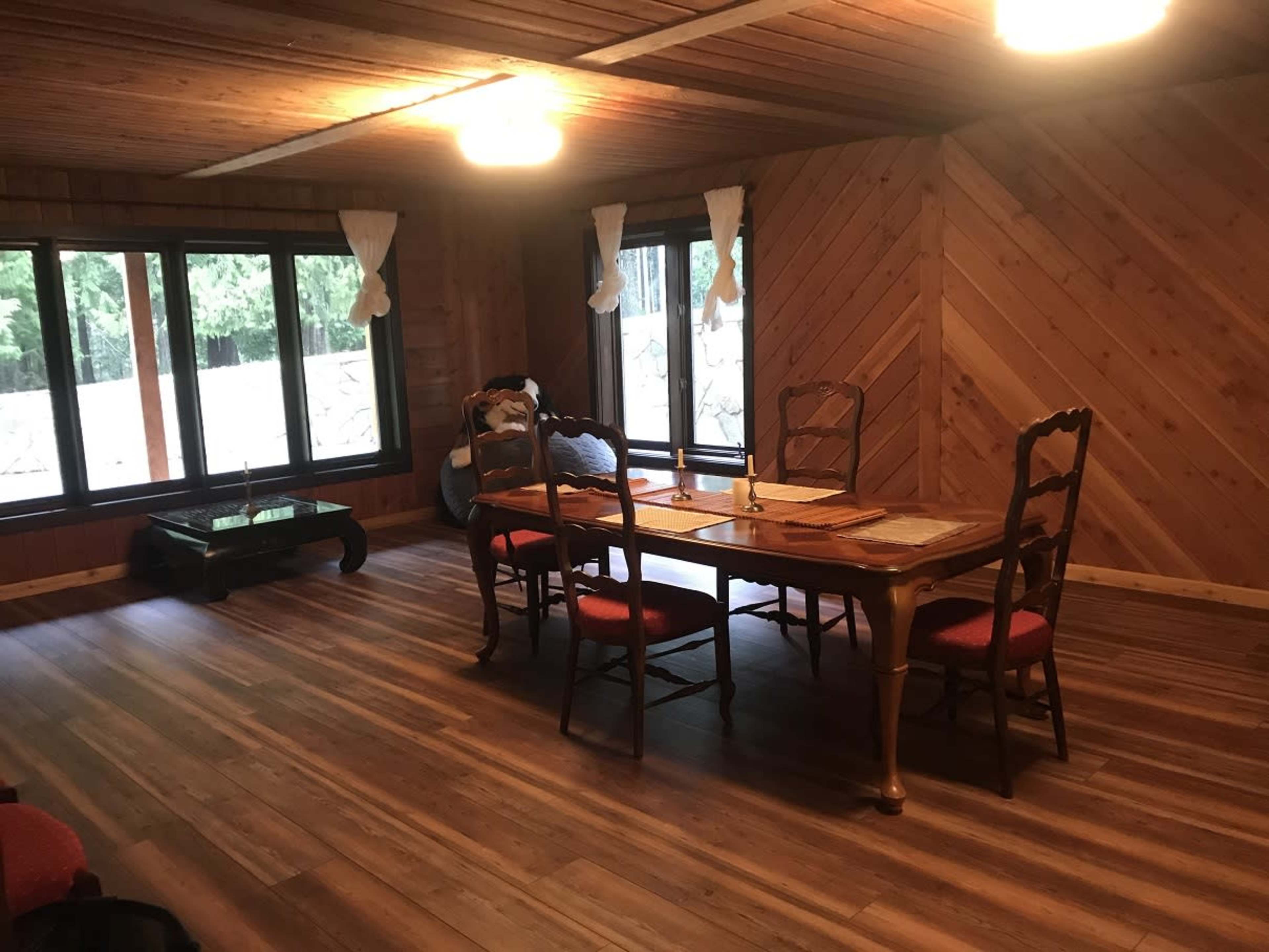 A wooden dining area features a table with chairs, large windows providing natural light, and a wooden floor.