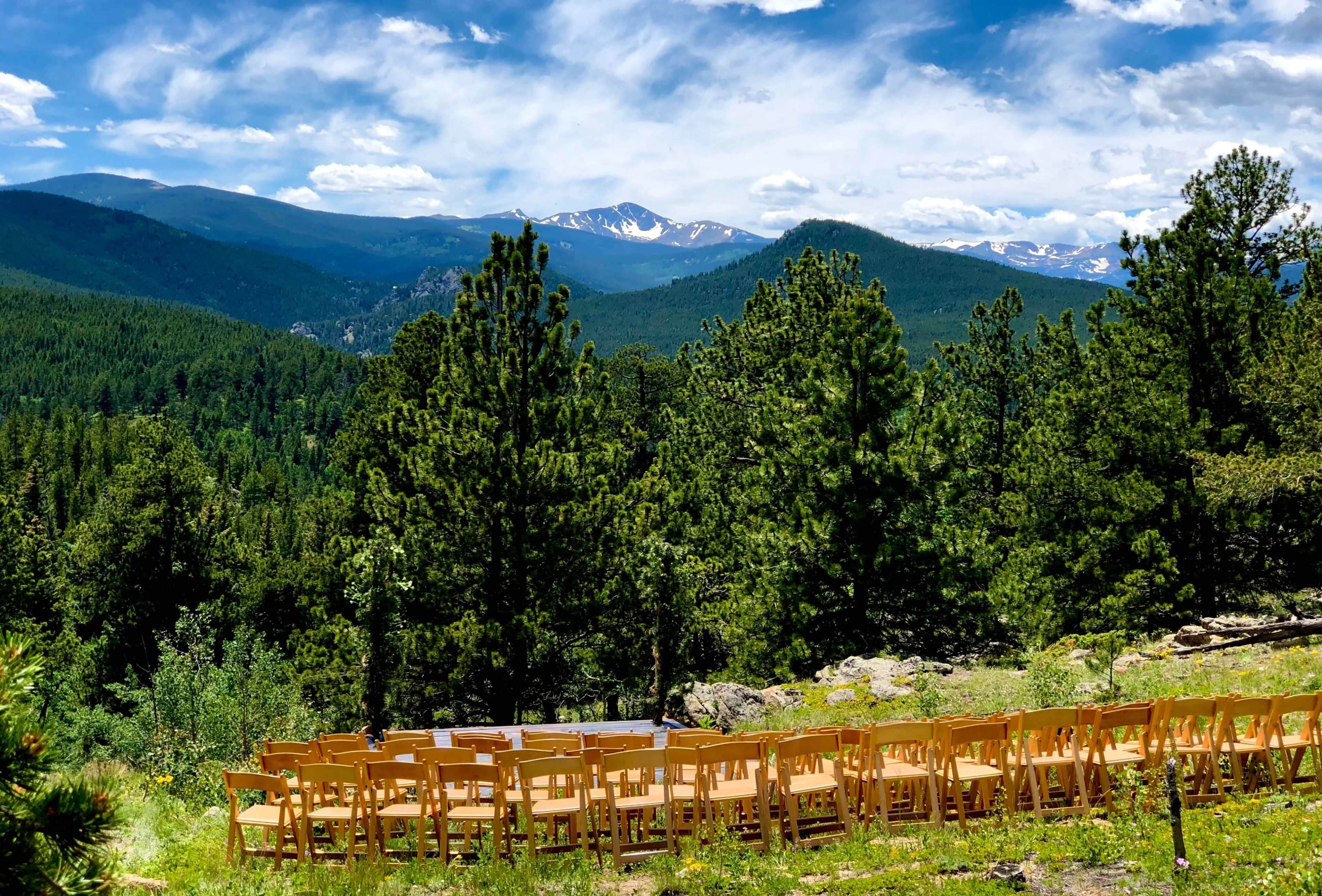 A row of orange wooden chairs is set up on a grassy area overlooking a mountainous landscape with trees and a clear sky.