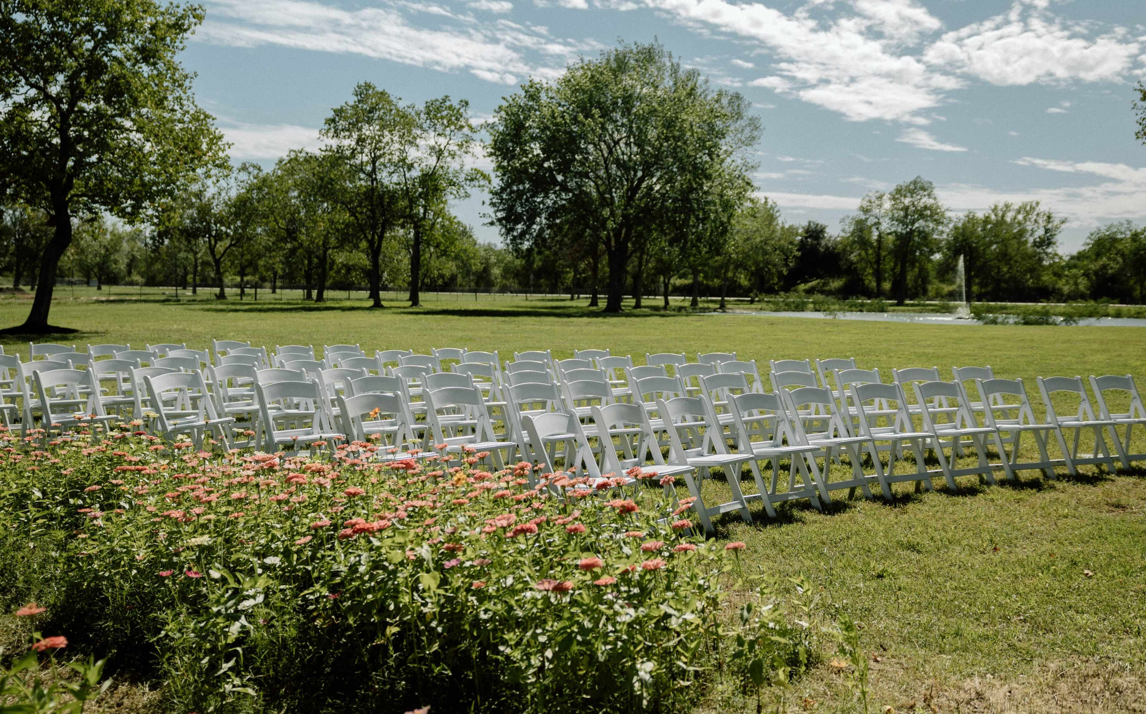 A row of white chairs is arranged on a grassy field near blooming flowers and trees under a clear sky.