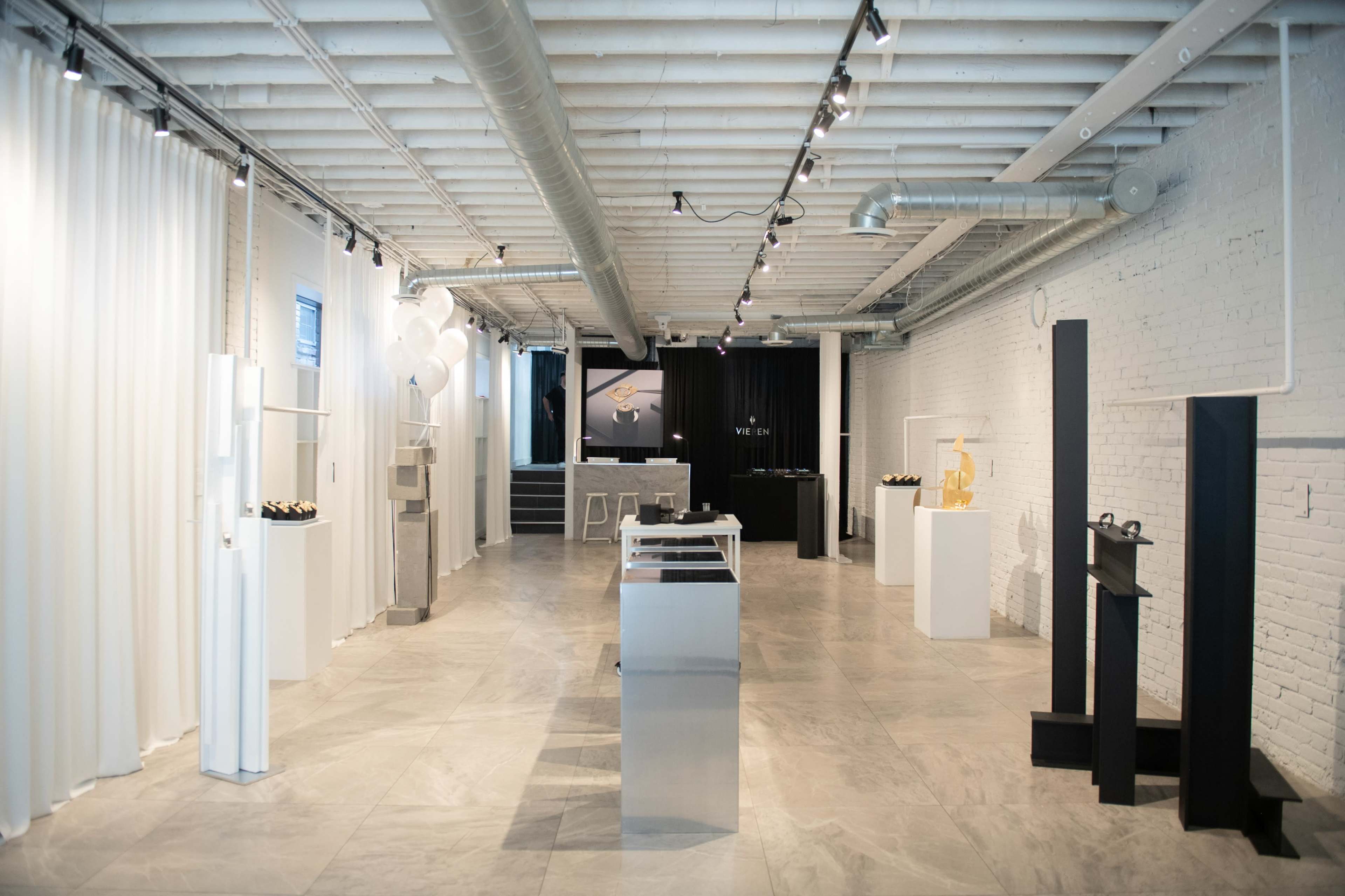 The image depicts a modern gallery space with white walls, polished stone flooring, and various artistic displays centered throughout the room.