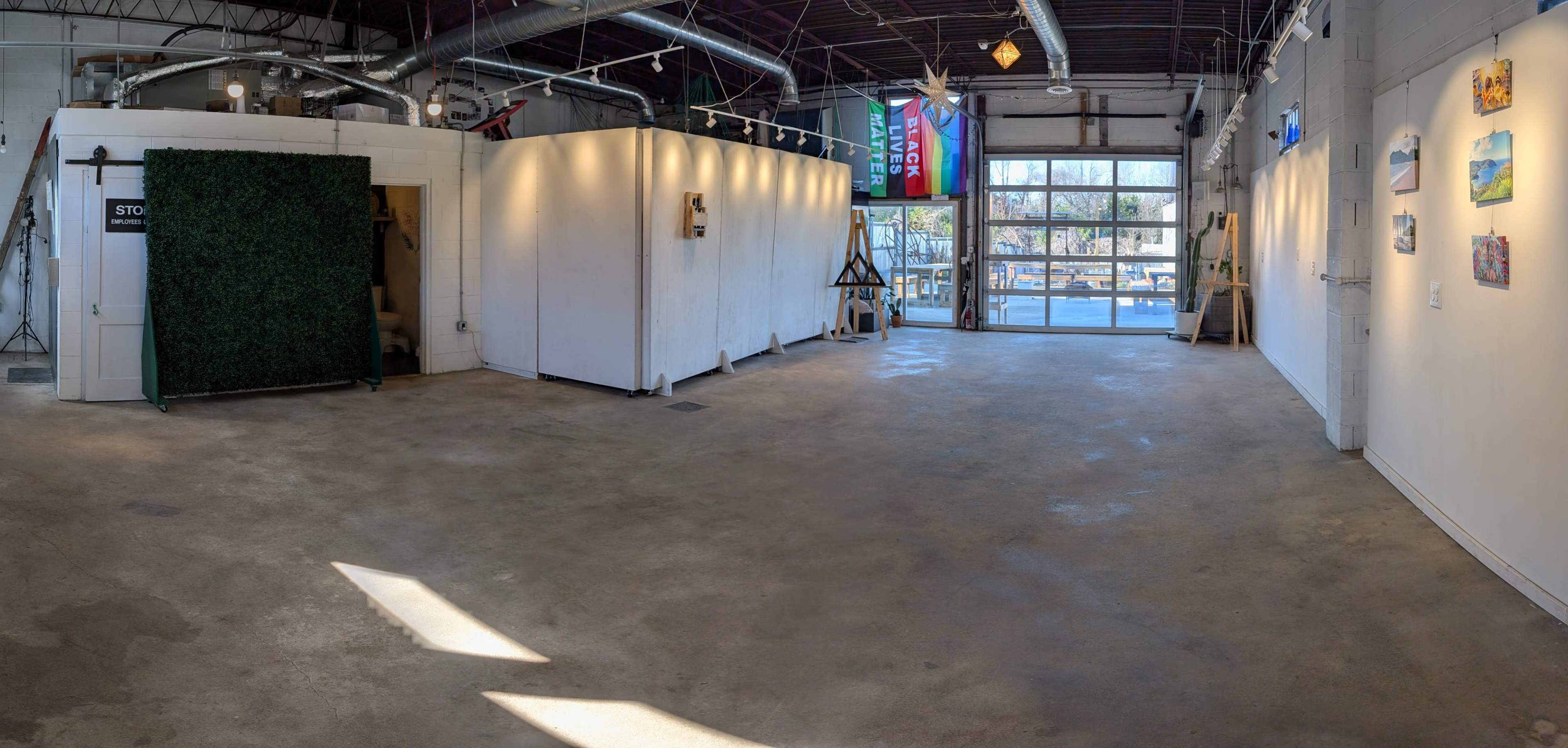 The image shows a spacious, well-lit interior of an art gallery or studio with white walls, a concrete floor, and large windows allowing natural light to enter.