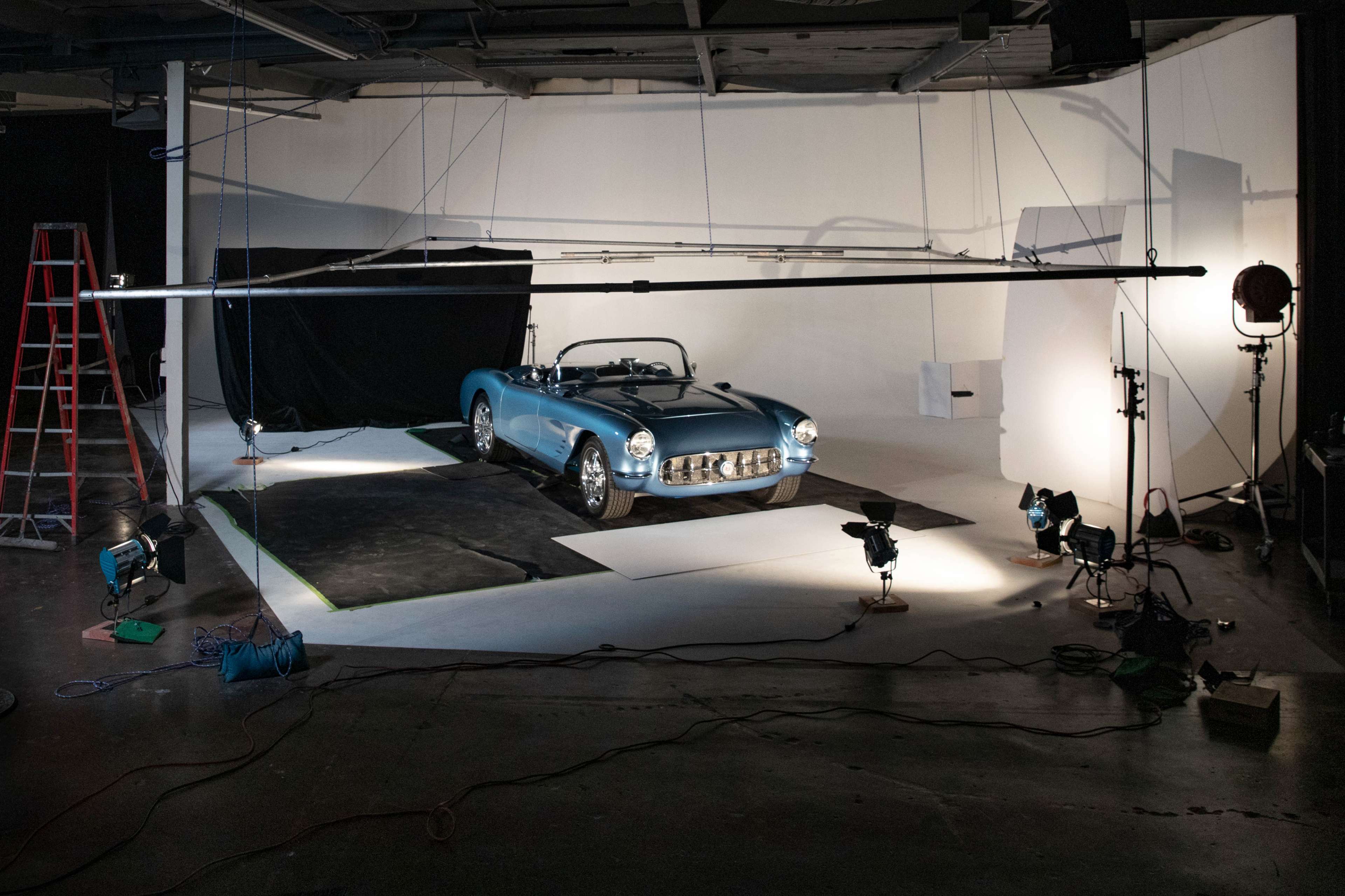 A classic blue convertible car is positioned on a photo studio set, surrounded by lighting equipment and a ladder.