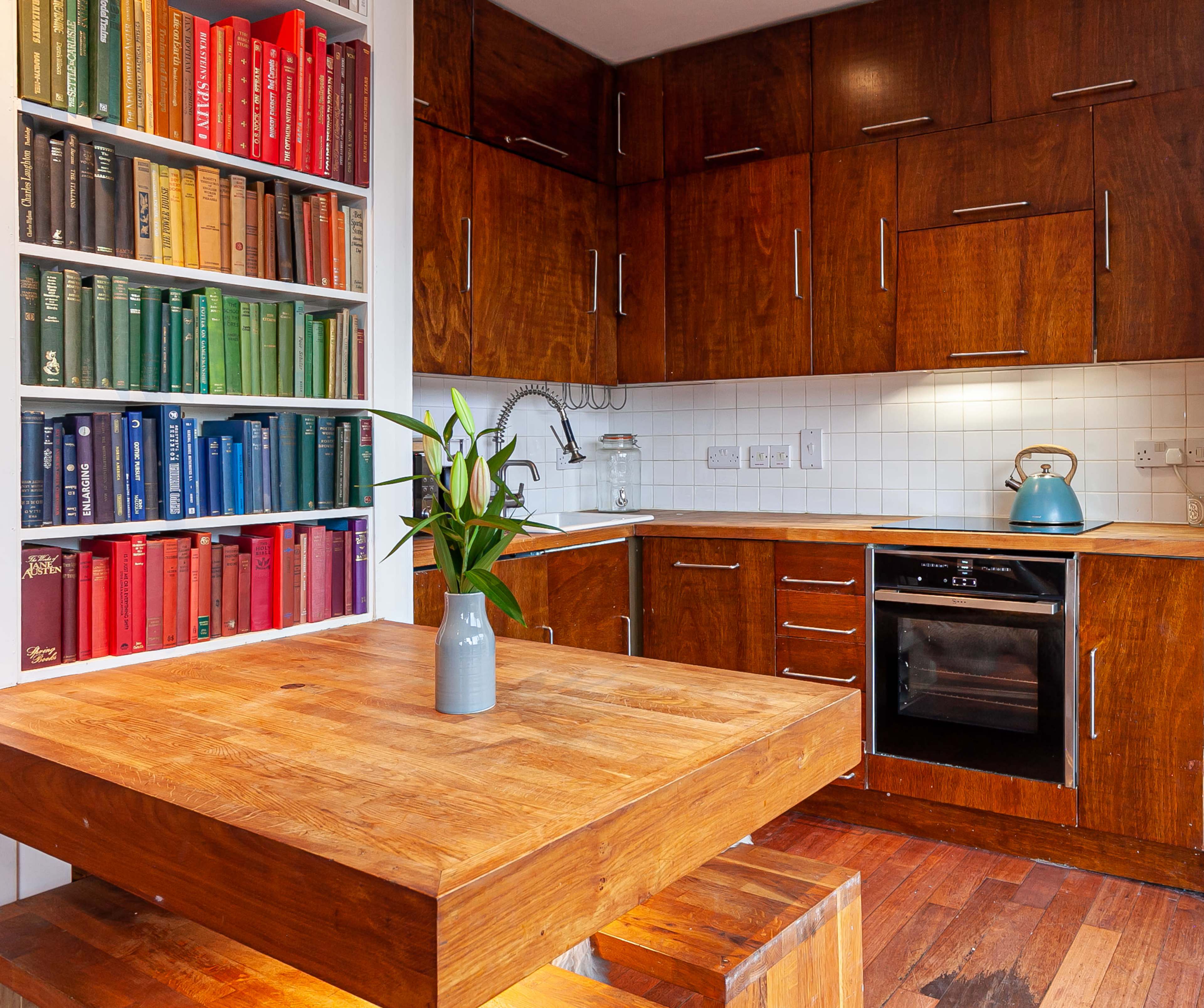 A modern kitchen with wooden cabinets, a colorful bookshelf filled with books, and a wooden dining table with a small vase of flowers.