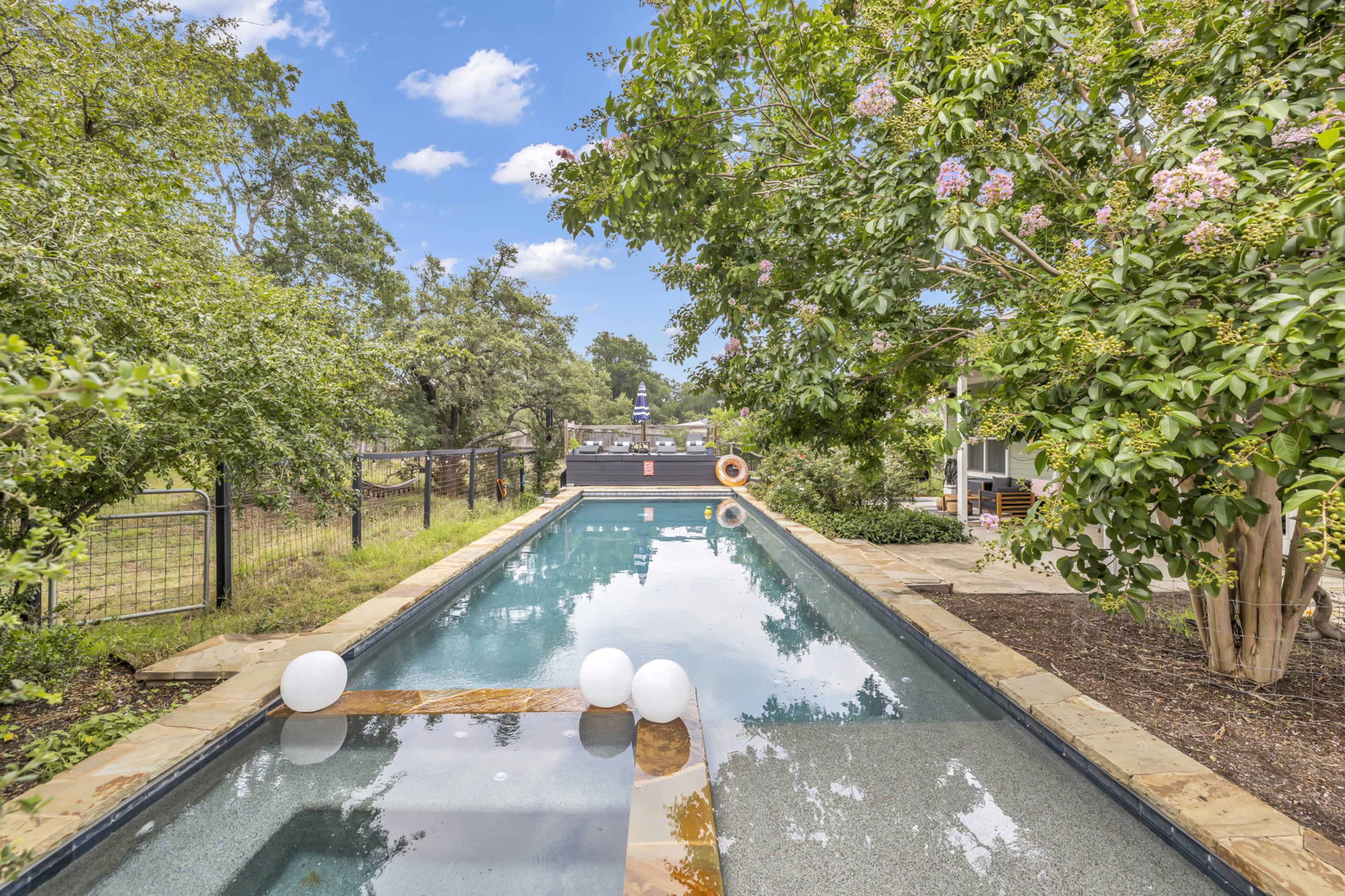 The image shows a long, narrow swimming pool surrounded by trees and landscaping, with a few decorative white orbs floating on the water's surface.