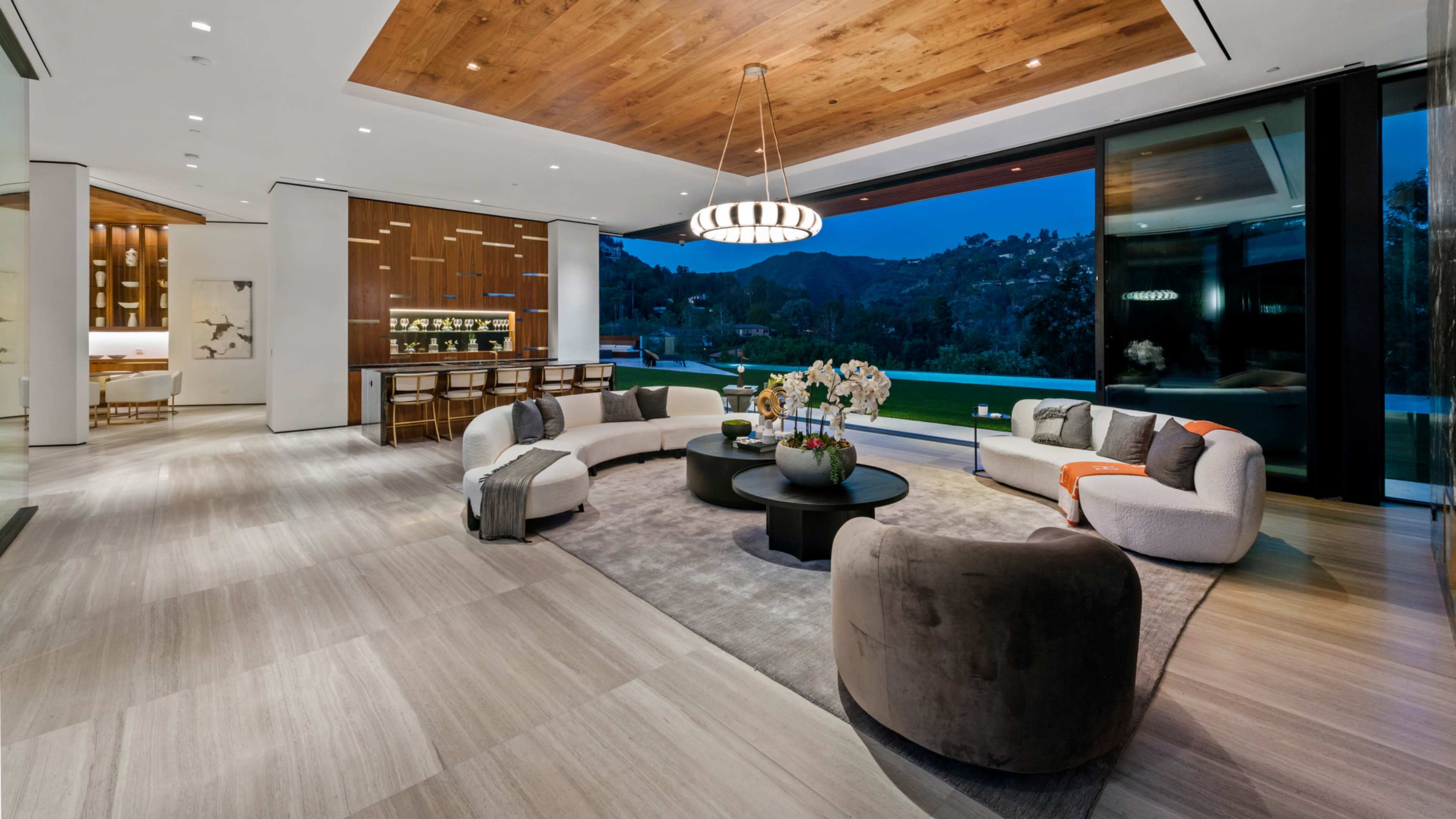 The image depicts a modern living room with a large glass wall providing a view of the outdoors, featuring a circular seating arrangement around a central coffee table and warm wooden accents on the ceiling.