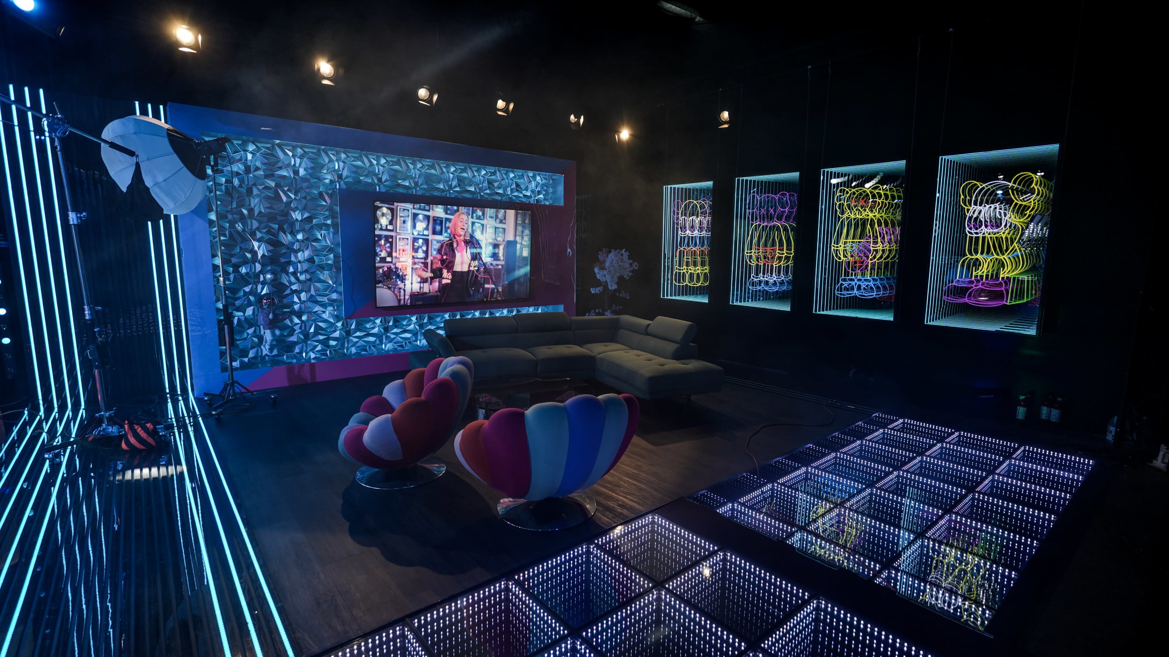 The image shows a modern TV studio set featuring colorful furniture, neon art, and a large screen displaying digital visuals.