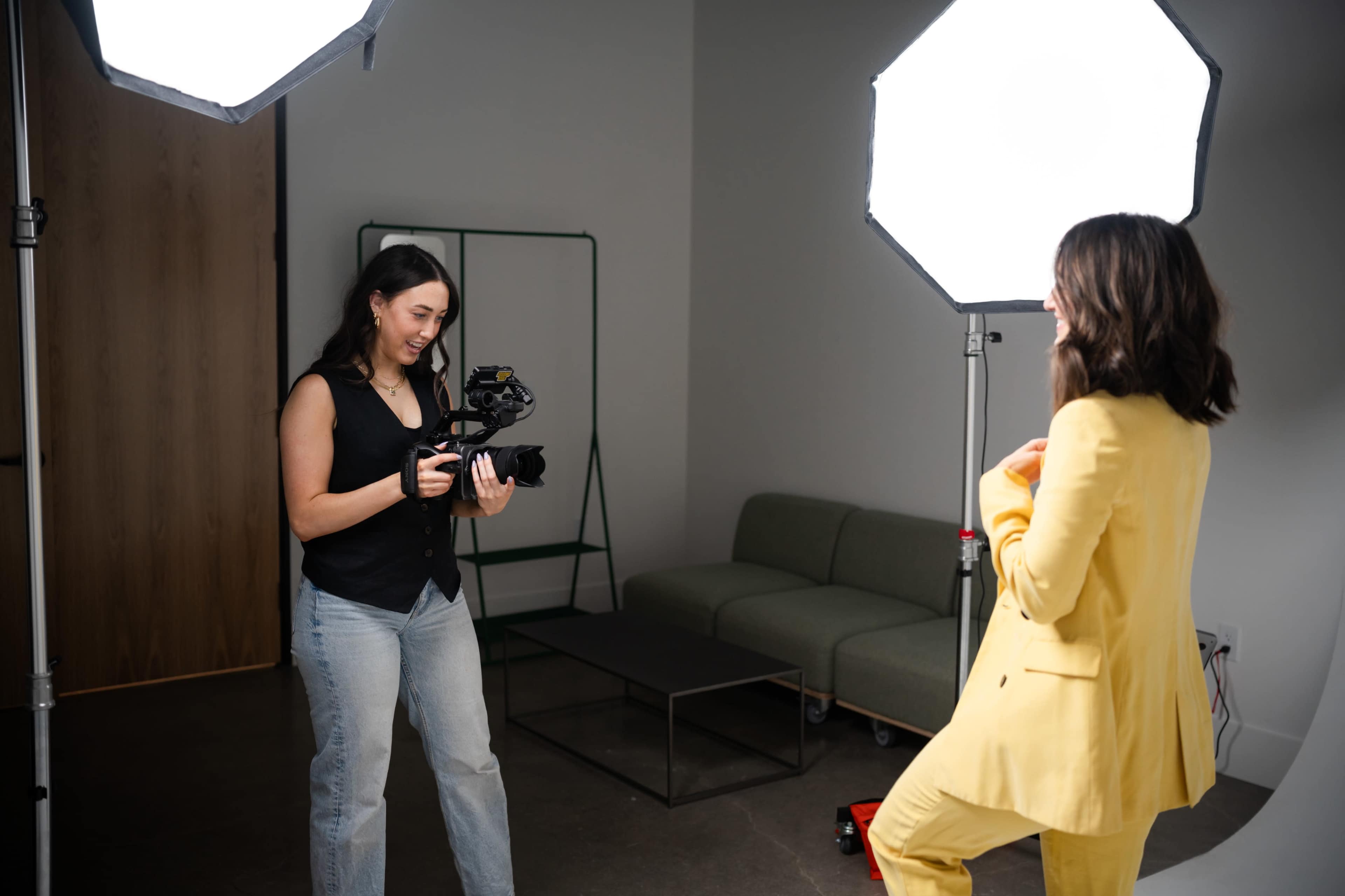 A woman in a yellow suit poses for a photo while another woman holds a camera in a well-lit studio setting.