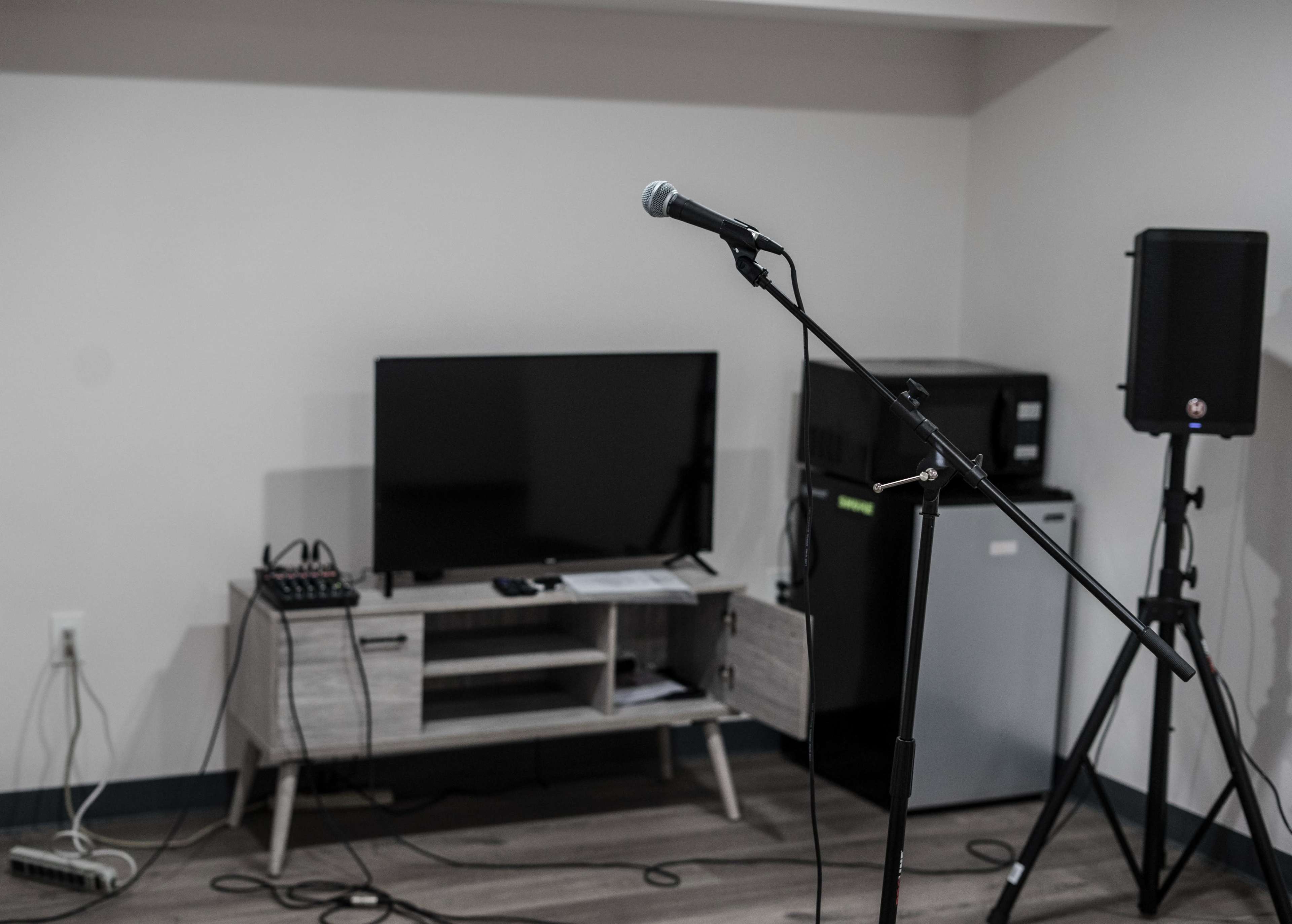 A microphone stands on a stand in front of a television and a small refrigerator in a minimalistic room.