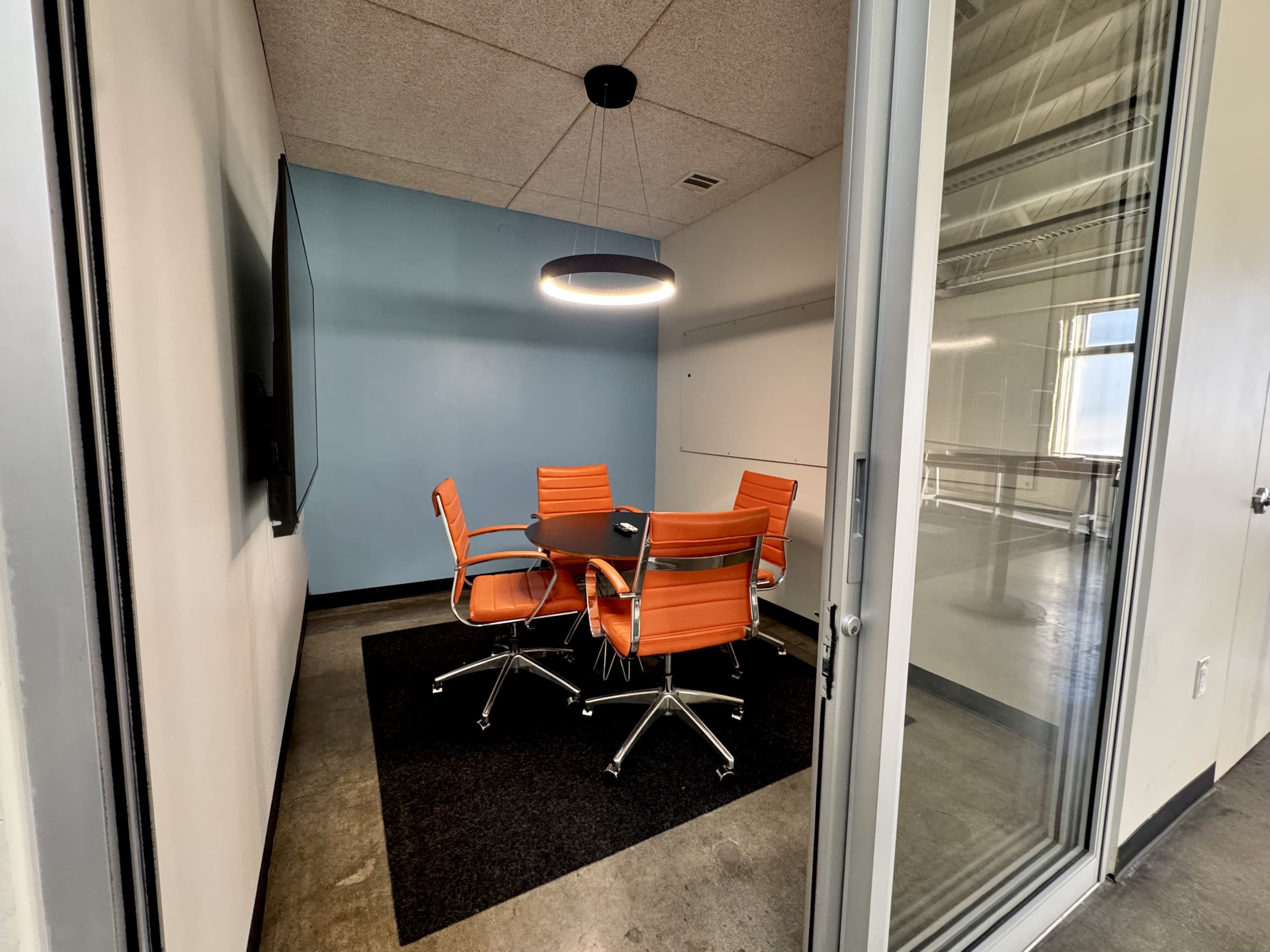 A glass-walled conference room features a round table surrounded by four orange chairs and is illuminated by a modern pendant light.