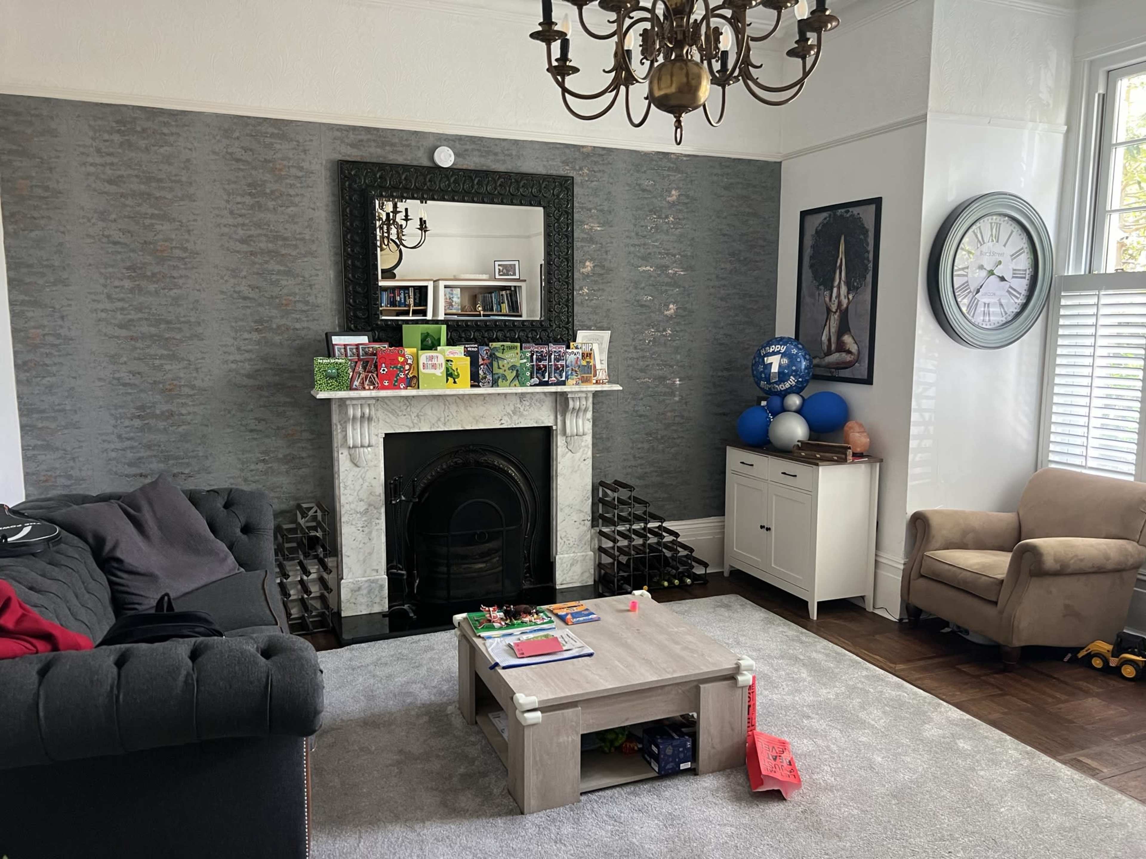 The image shows a living room with a dark couch, a coffee table cluttered with toys, a decorative fireplace with a large mirror above it, and various items on a shelf along with a blue globe and a wall clock.