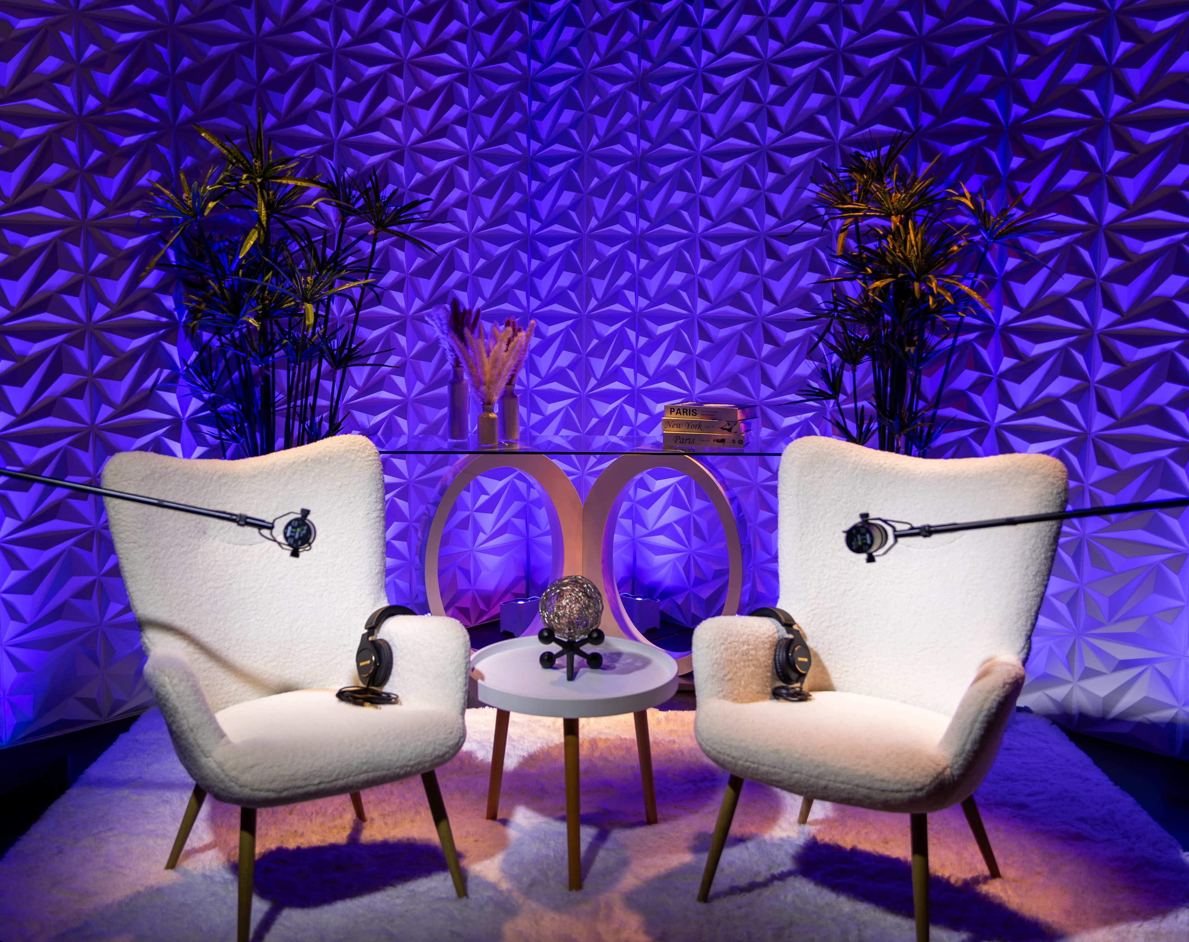 The image features a cozy podcasting setup with two plush chairs, a small table, and a decorative backdrop, all lit with purple lighting.