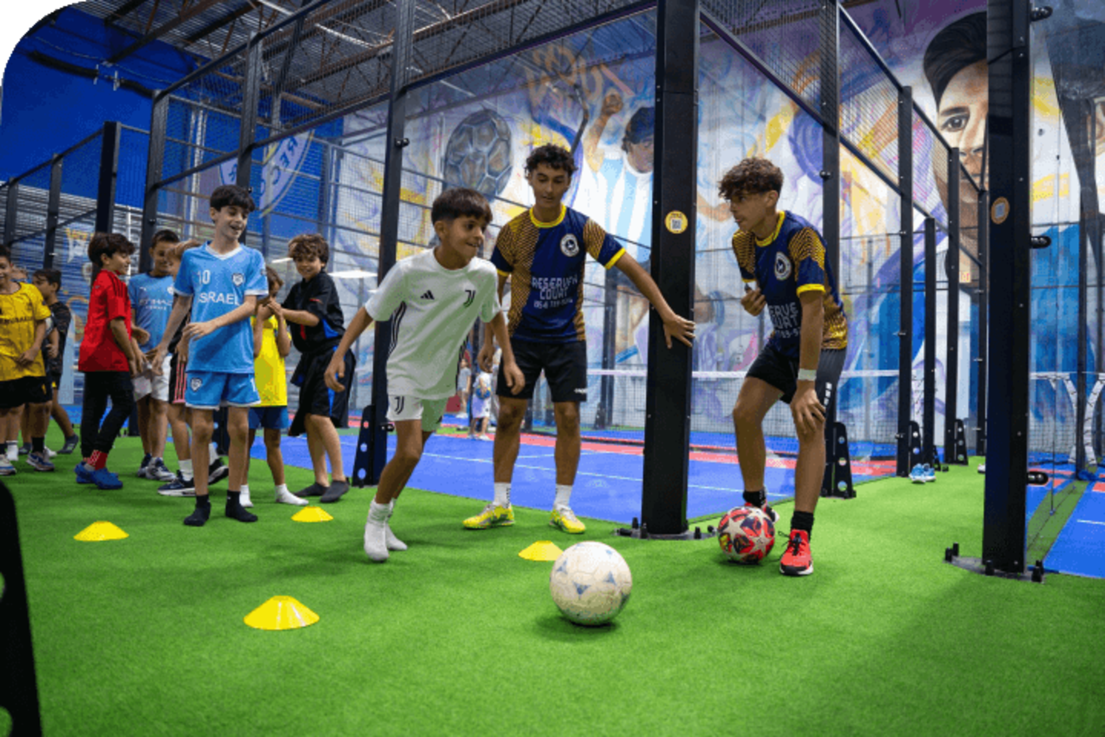 Children are participating in a soccer training session on an indoor field, navigating cones and dribbling balls while others observe.