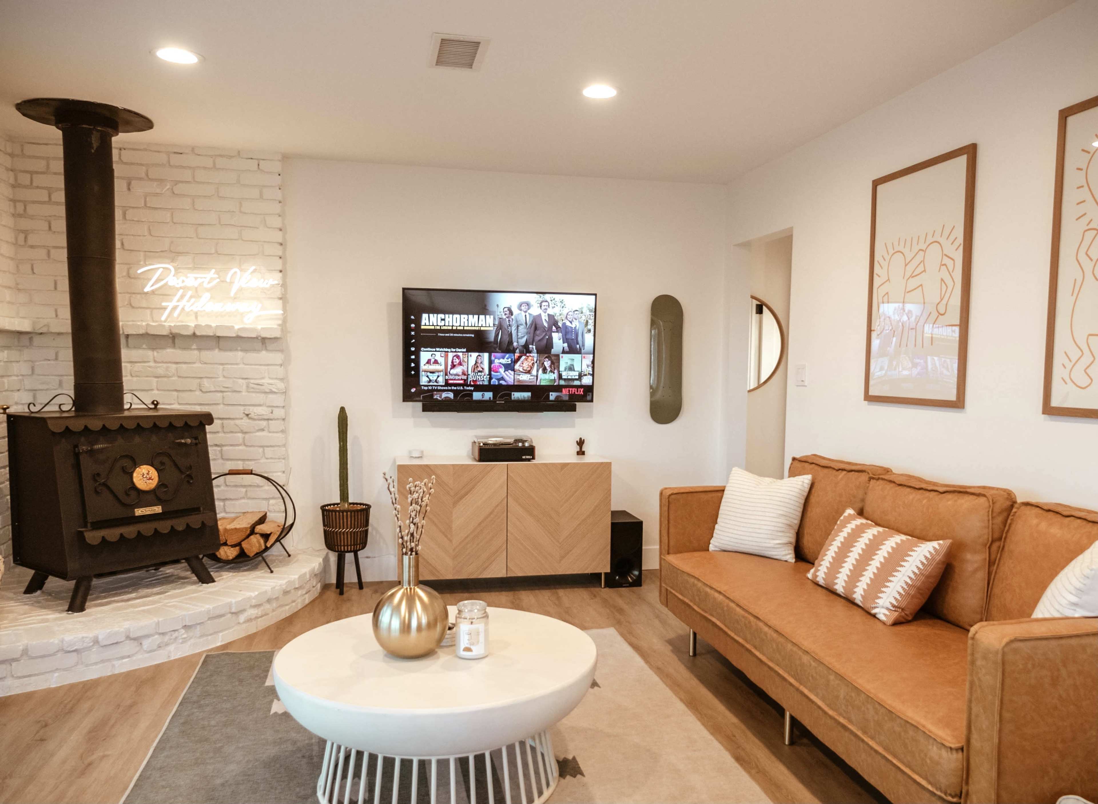 The image shows a modern living room with a brown sofa, a round coffee table, a television mounted on the wall, and a black metal fireplace.