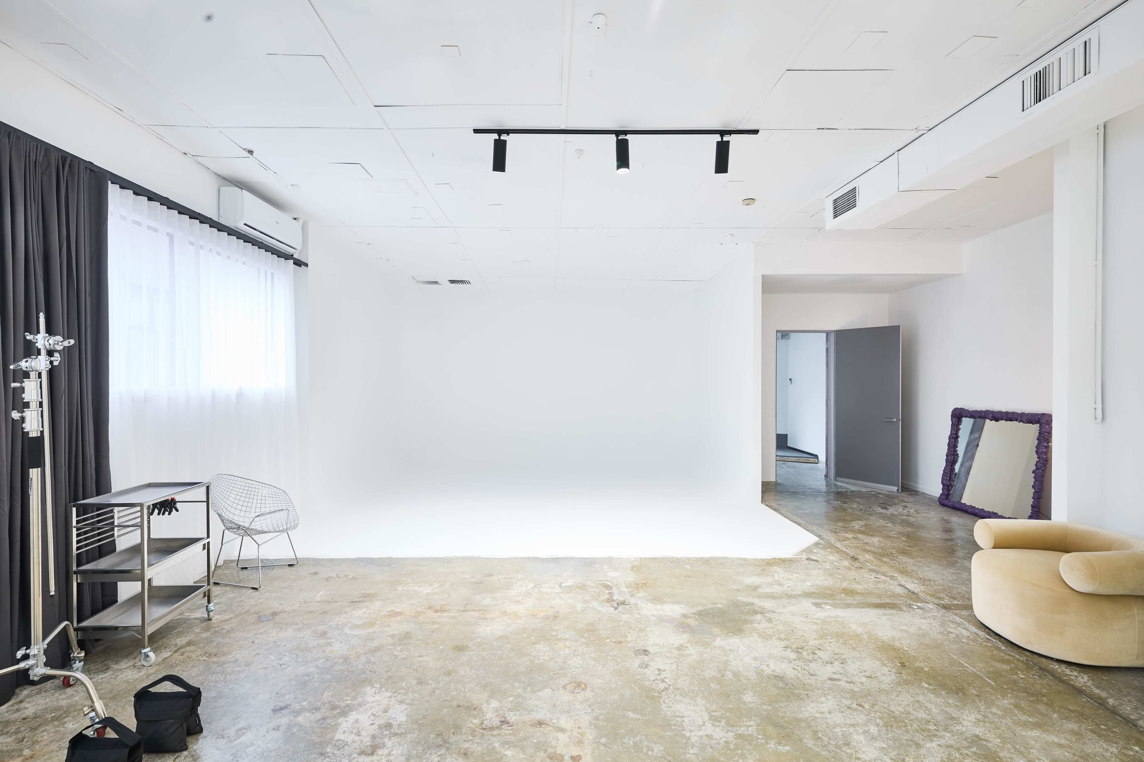 A spacious, well-lit studio features a white backdrop, concrete flooring, and minimal furnishings including a chair and a large mirror.