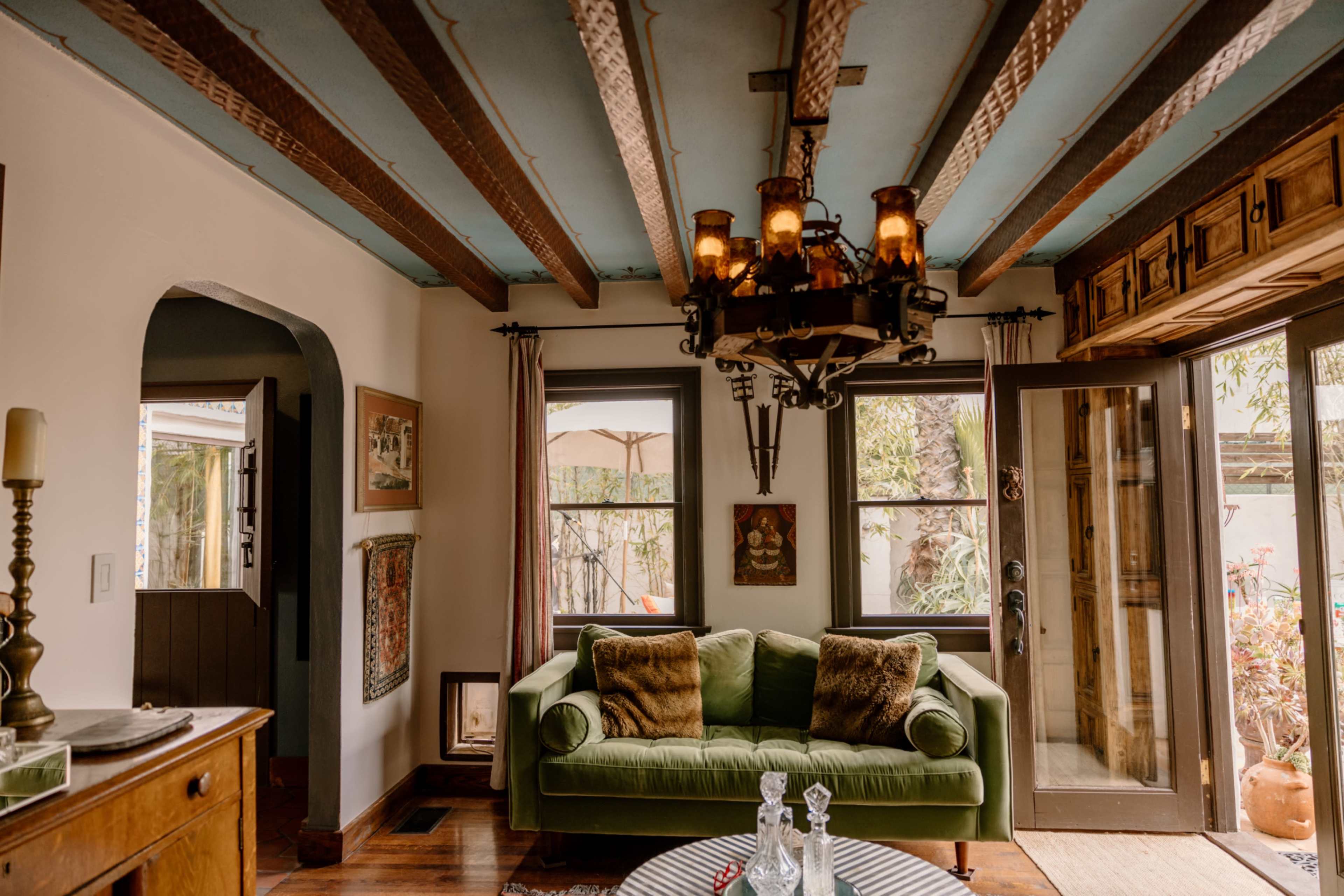 A cozy living room features a green couch, wooden beams overhead, and a chandelier, with large windows providing a view of an outdoor area.