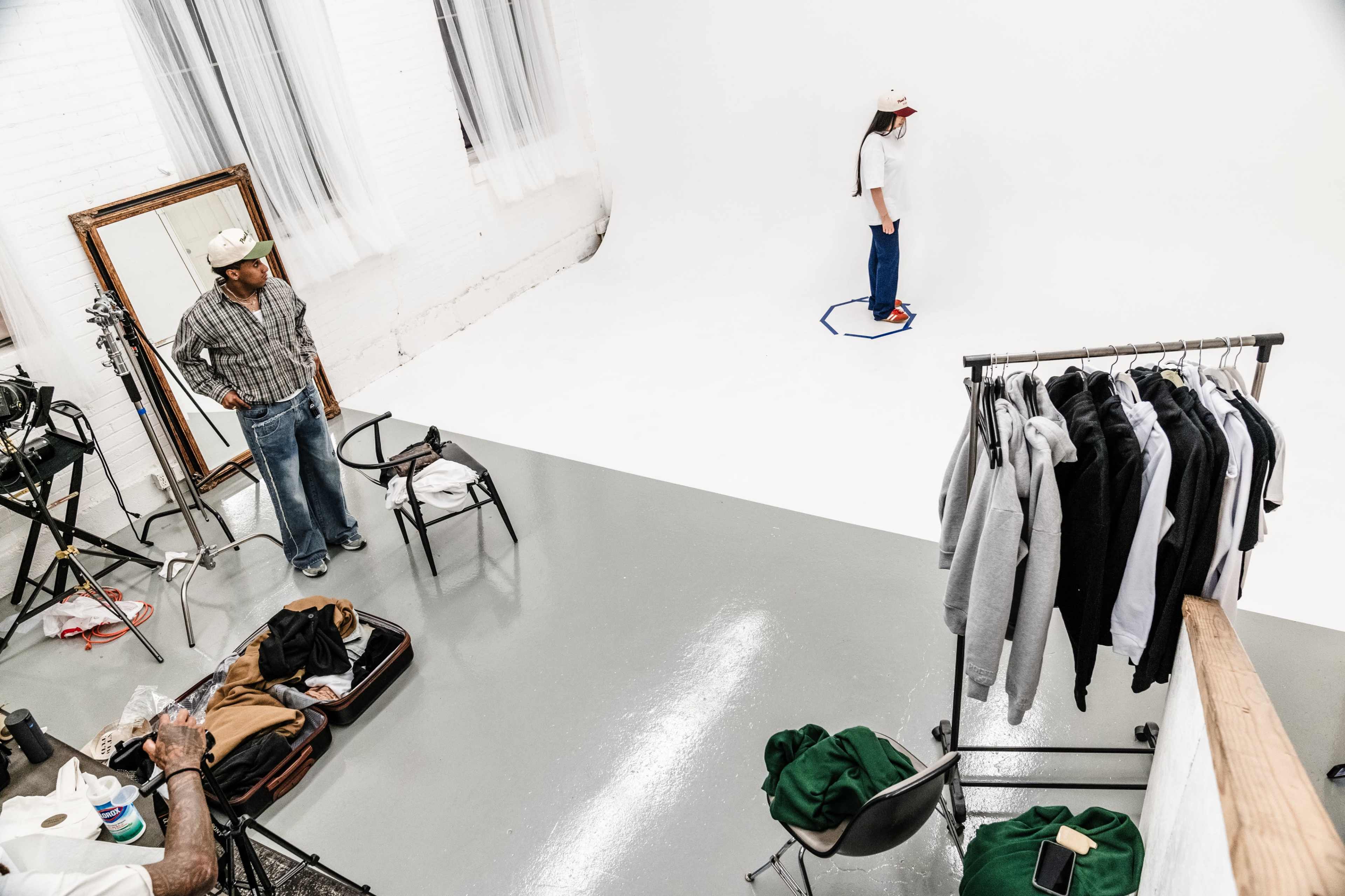 A model stands inside a blue hexagonal mark on a blank white backdrop while a photographer works in a studio filled with clothing on hangers and equipment.