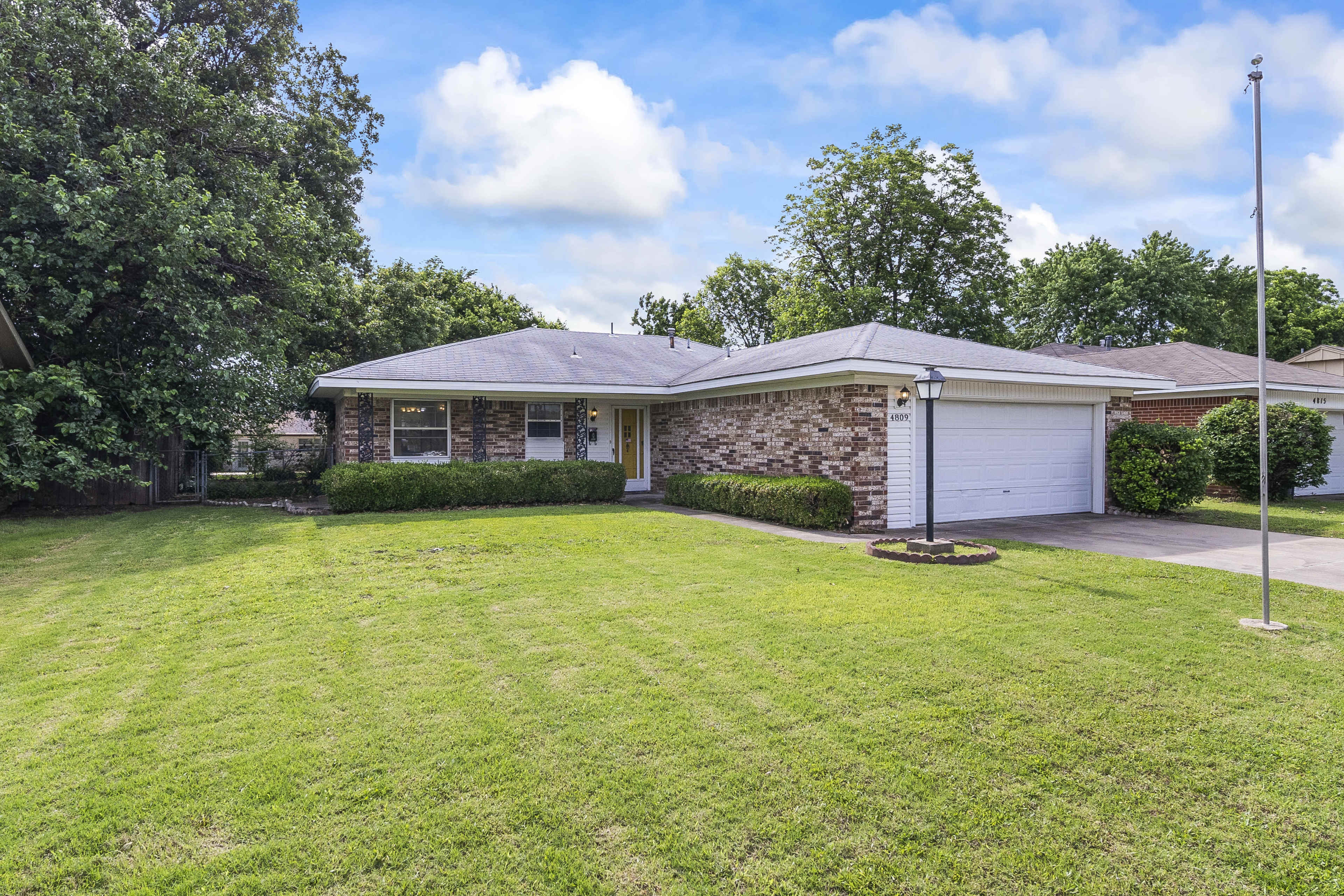 A single-story brick house with a garage, situated on a neatly maintained lawn and surrounded by trees and shrubs.