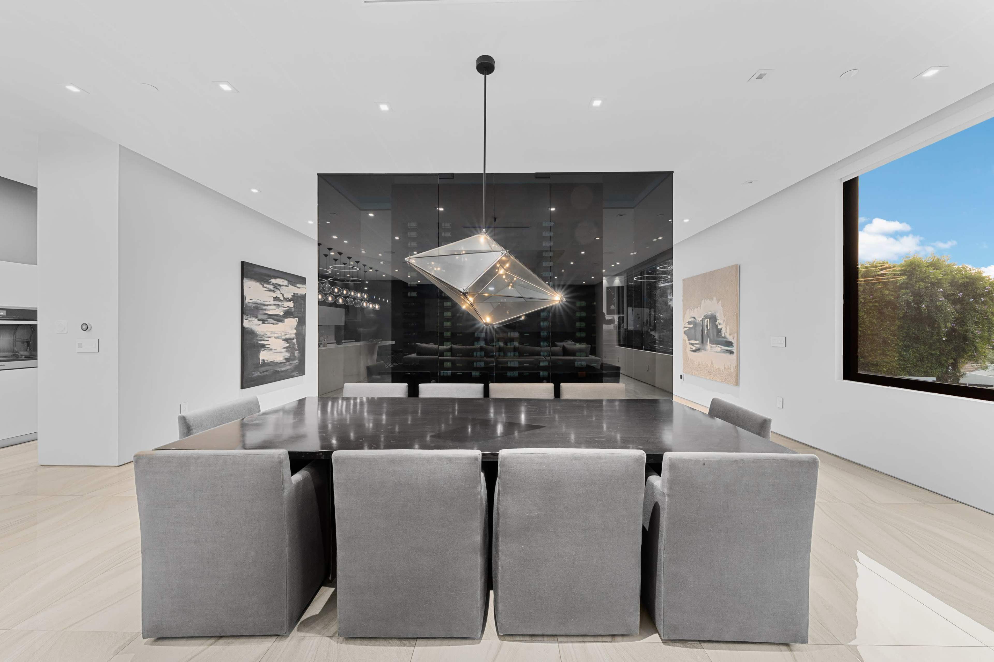 A modern dining room features a large rectangular table surrounded by gray upholstered chairs, illuminated by a geometric hanging light fixture, with artworks on the walls and a glass display in the background.