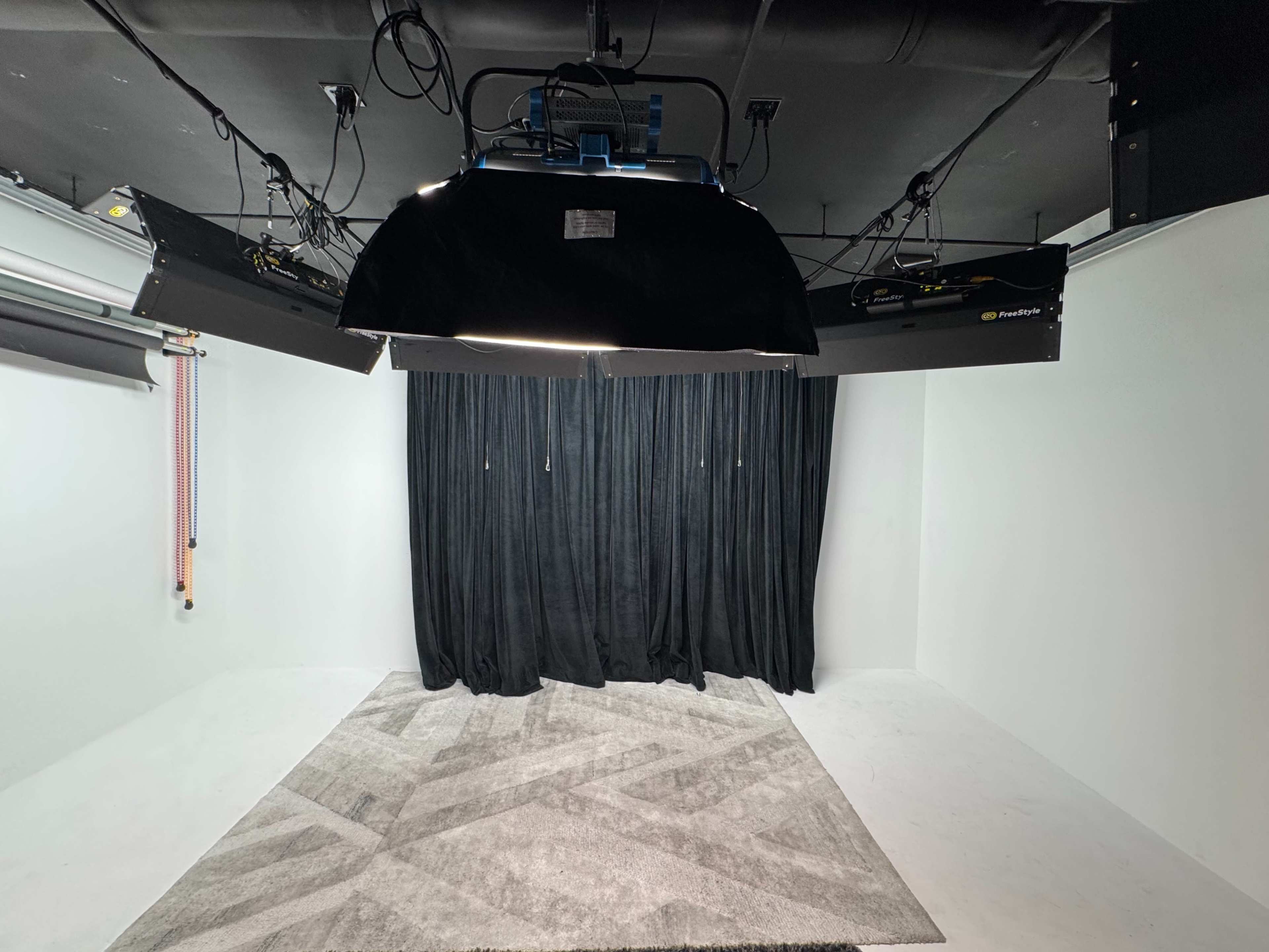 The image shows a photography studio with black backdrop curtains, overhead lights, and a gray area rug on the floor.