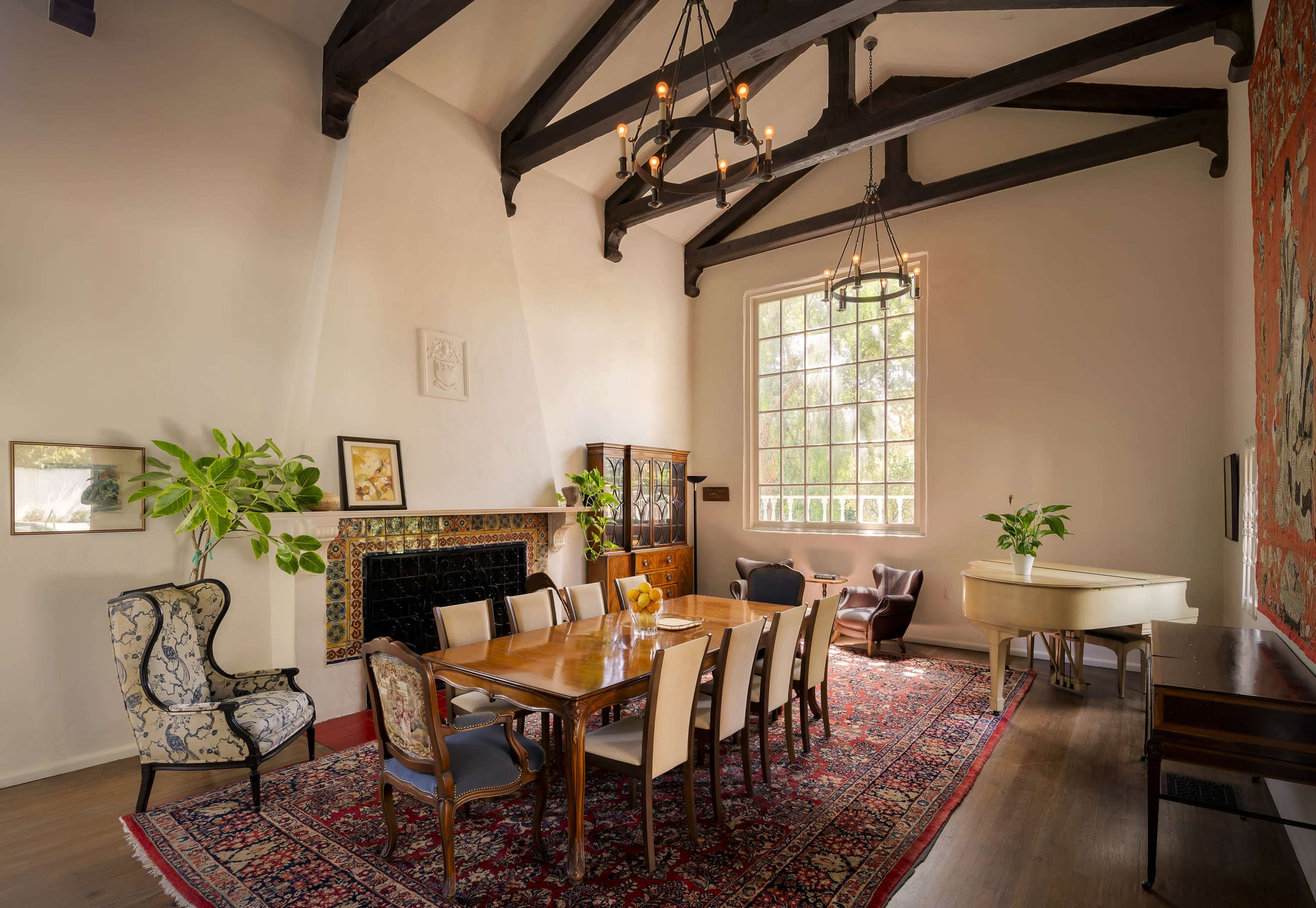 A spacious dining room features a long wooden table surrounded by chairs, with large windows and a high ceiling supported by wooden beams.