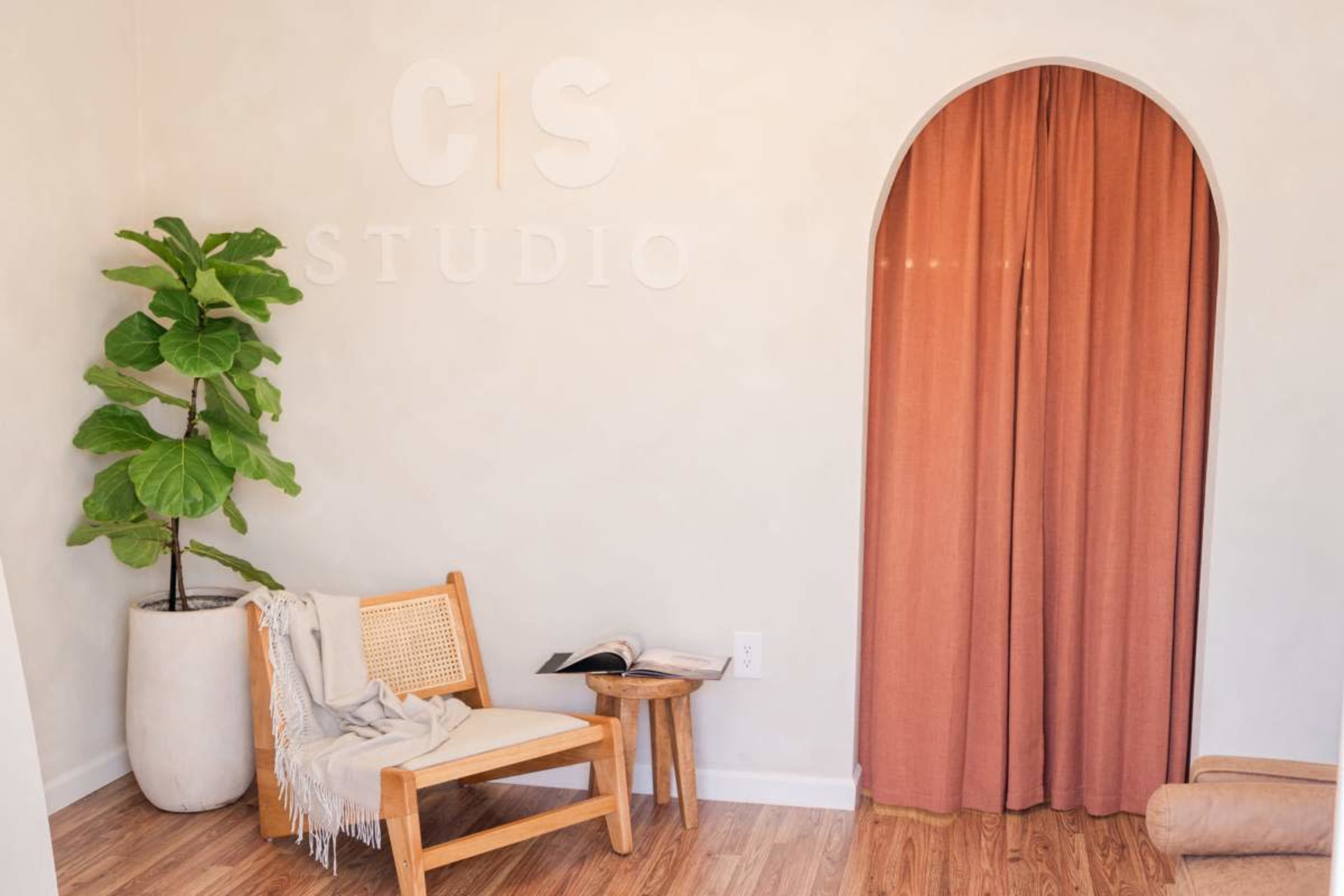 The image shows a cozy interior space featuring a small wooden chair, a draped curtain in an archway, a potted plant, and a partially opened book on the floor.