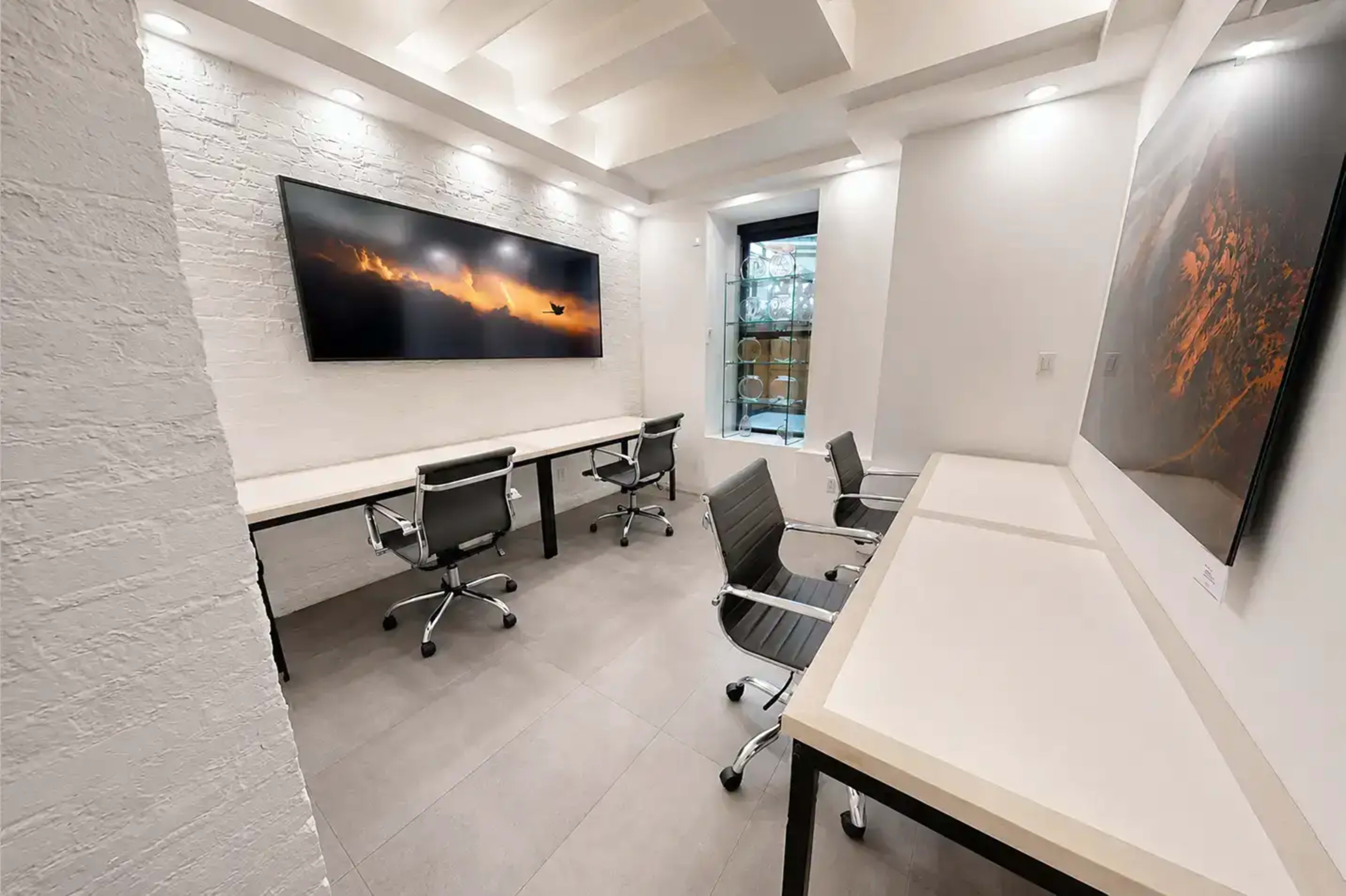 The image shows a modern office space with two desks, ergonomic chairs, a large wall-mounted screen displaying a scenic image, and bright overhead lighting.