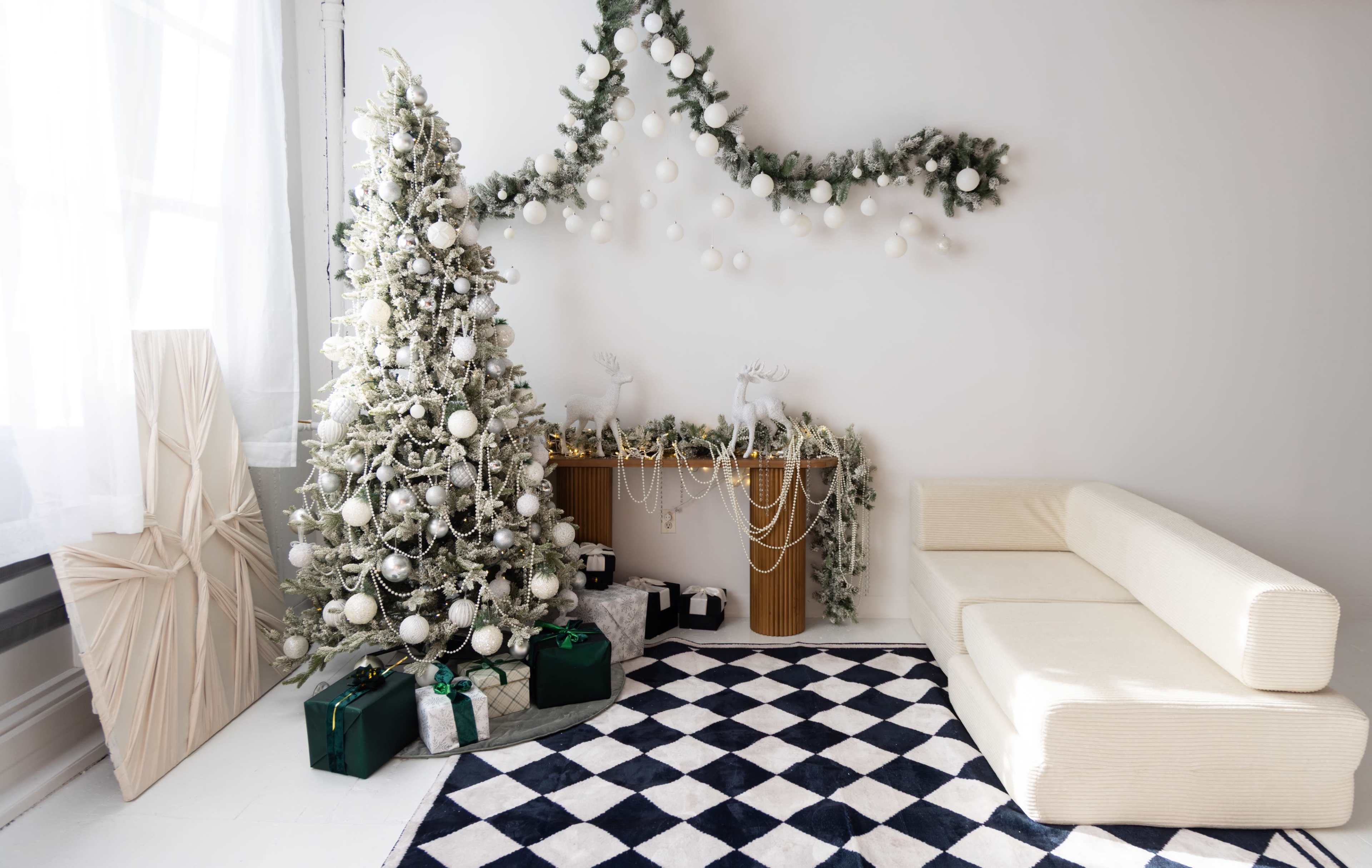 The scene features a decorated Christmas tree surrounded by gifts, a white sofa, and a wall adorned with garlands and ornaments in a bright, minimalist room.