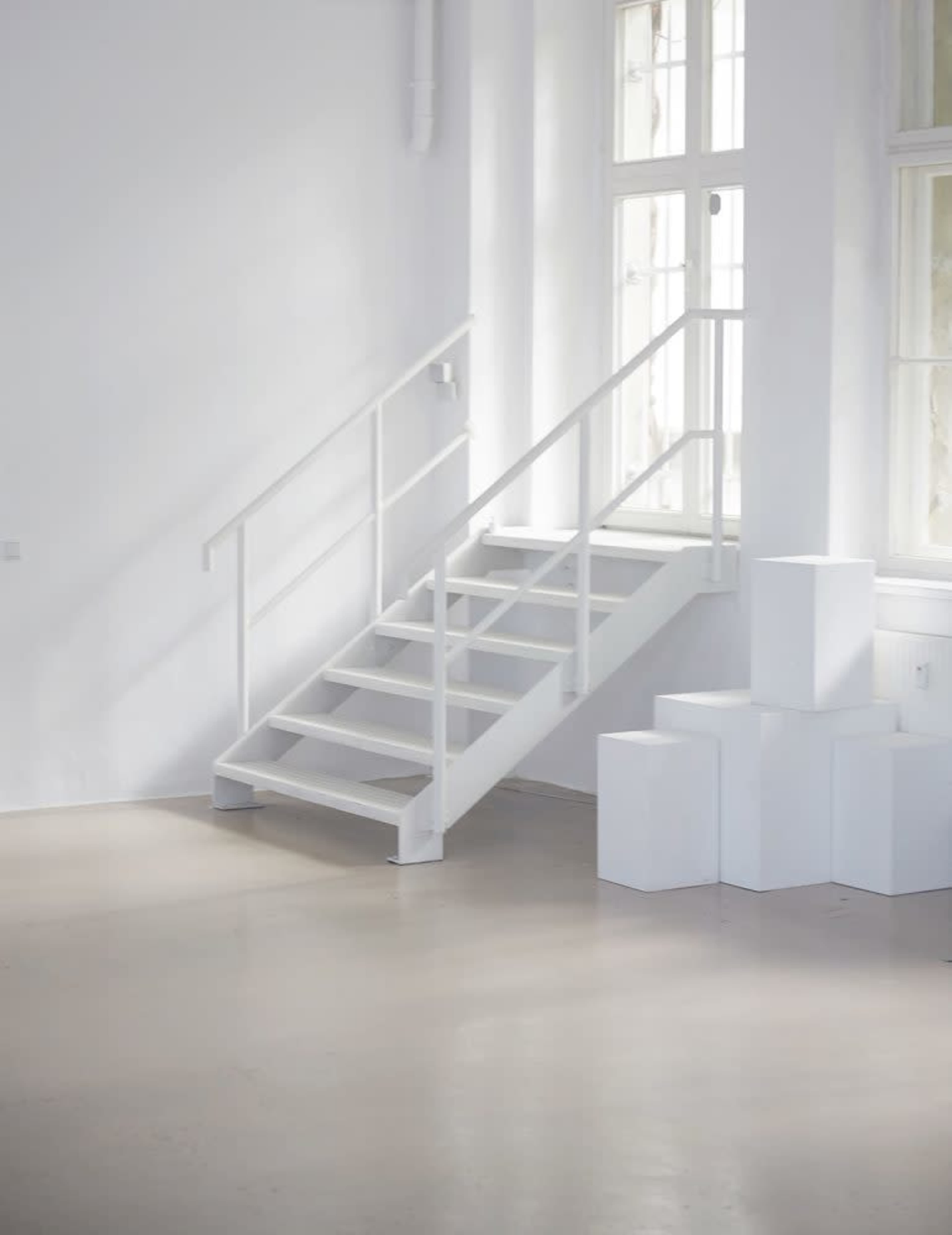 The image shows a minimalist, white interior space with a staircase and white cubes arranged on the floor.