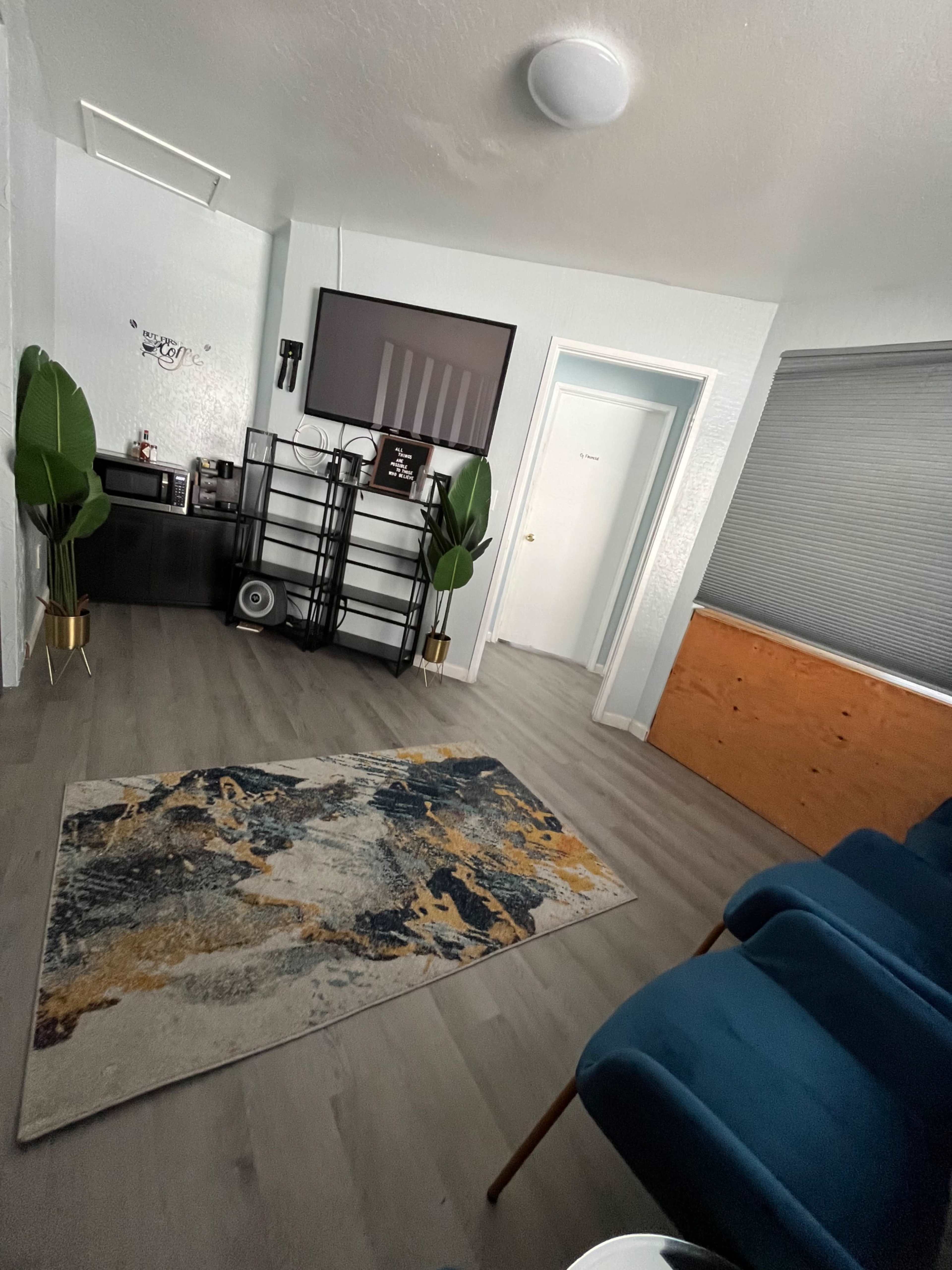 A neatly arranged living space features a large television on the wall, a shelving unit, a rug with an abstract design, and a pair of blue chairs.