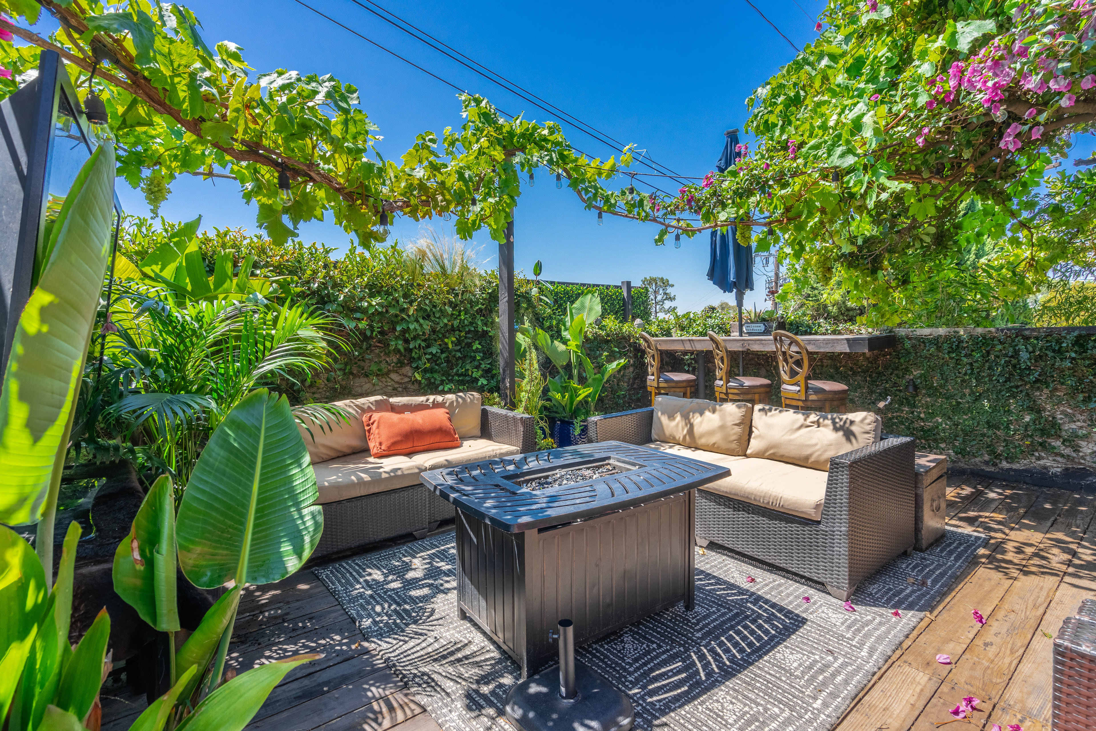 A patio area featuring a seating arrangement with cushions, a fire pit, and lush greenery overhead.