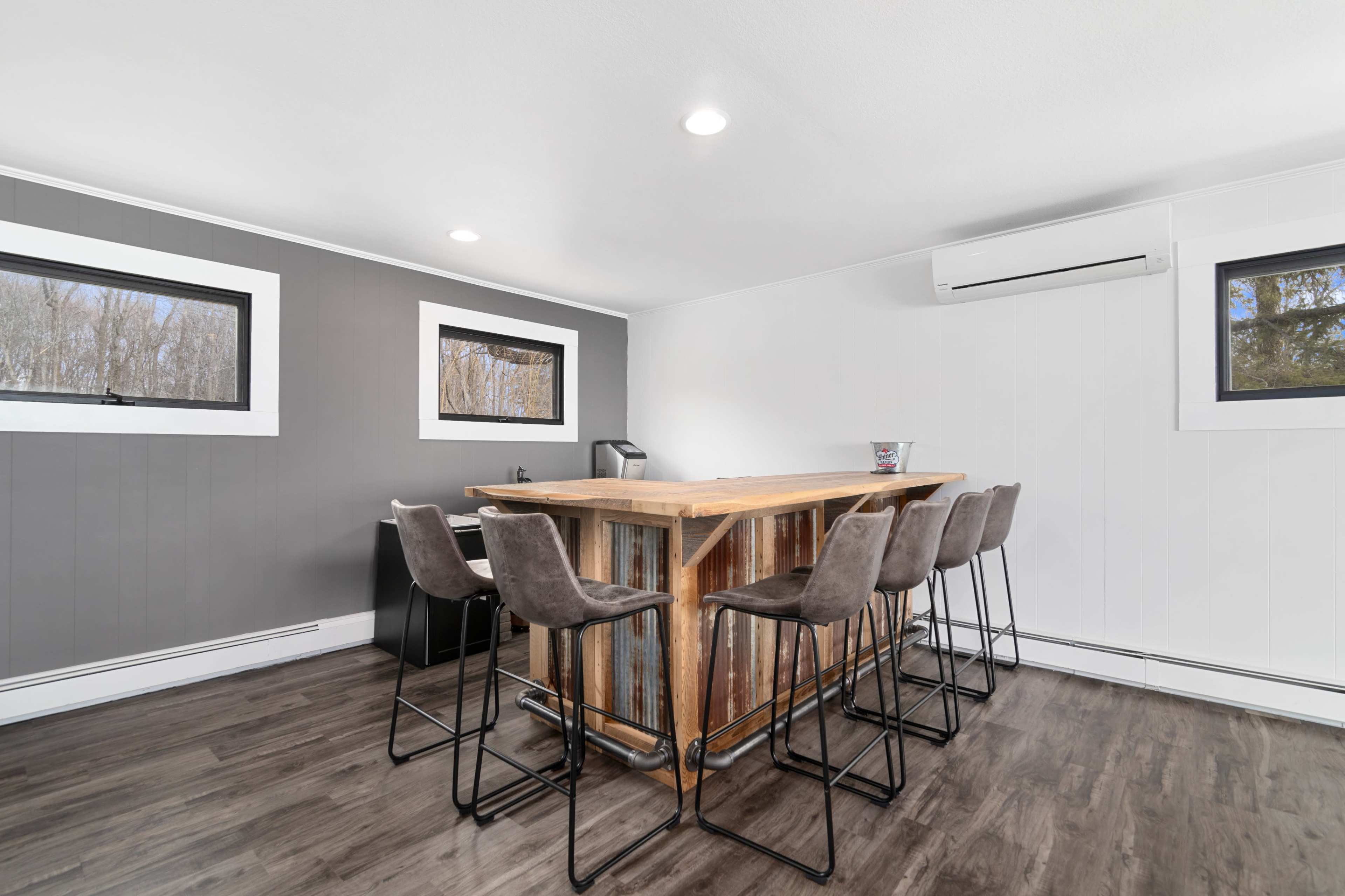 A modern interior space featuring a wooden bar with high stools, set against gray and white walls, and two windows that provide natural light.