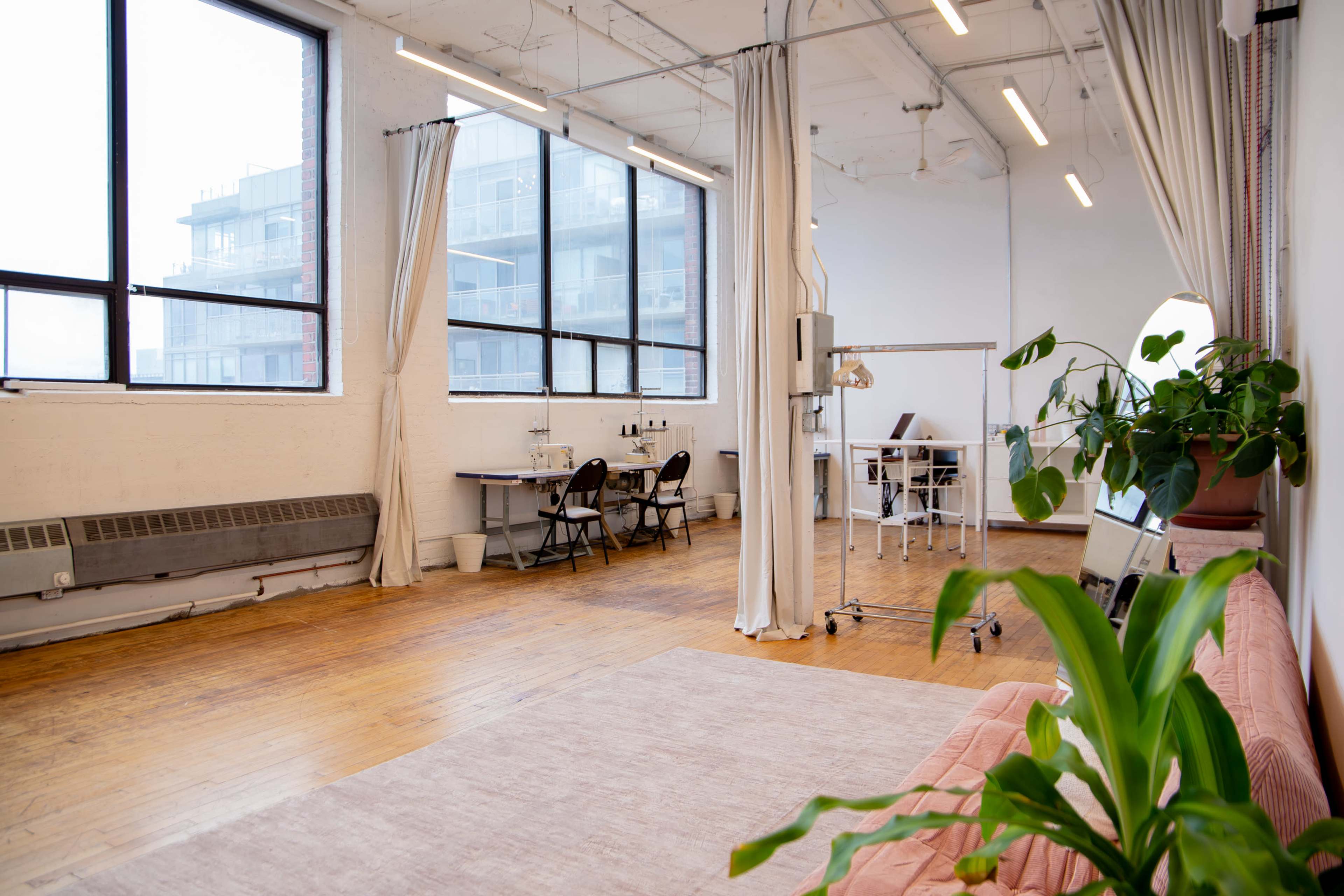 The image shows a spacious, brightly lit room with large windows, wooden flooring, and minimal furnishings, including a sofa, tables, and plants.
