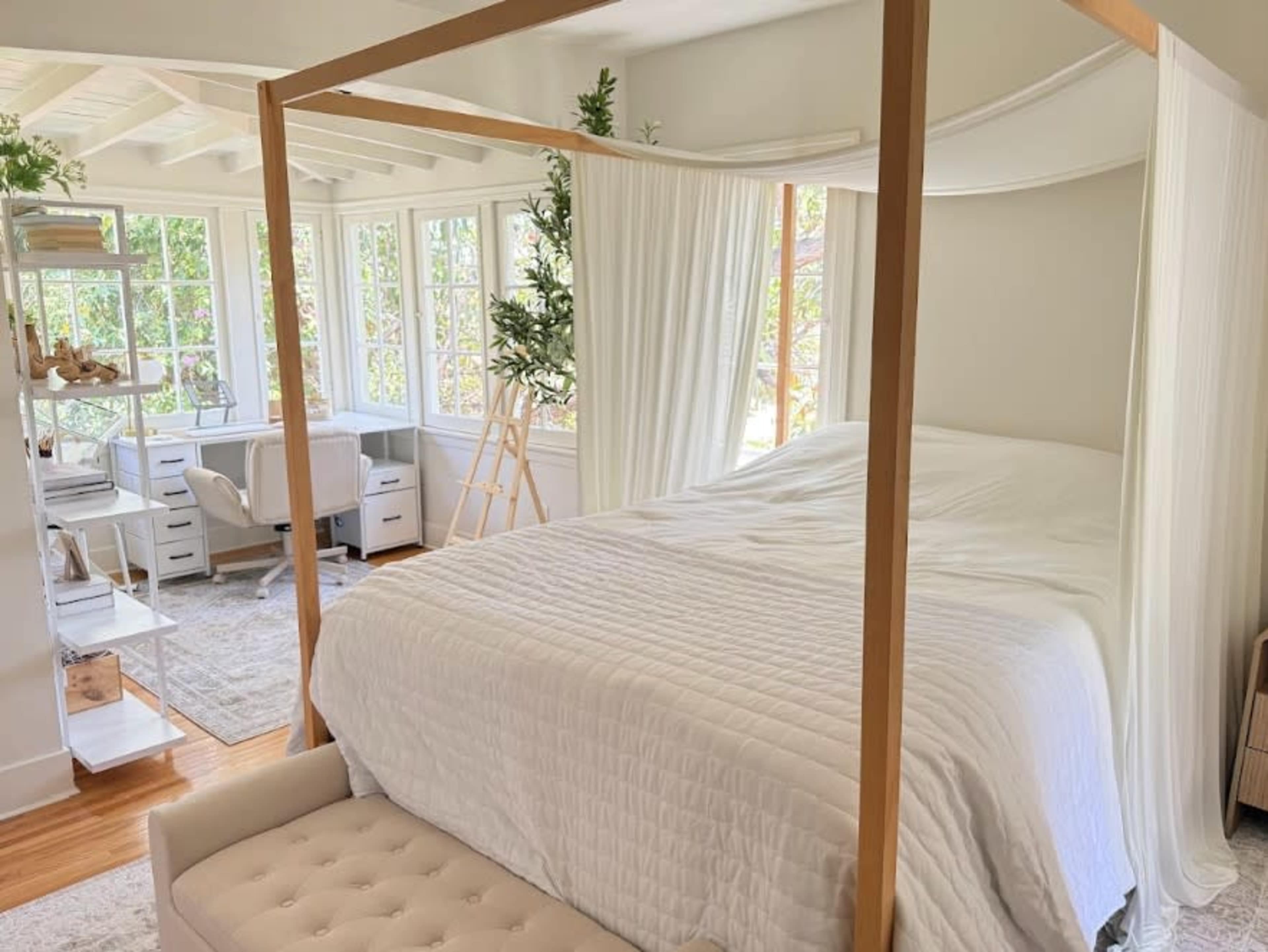 A light-filled bedroom features a four-poster bed draped with curtains, paired with a cozy sitting area and a workspace by the windows.