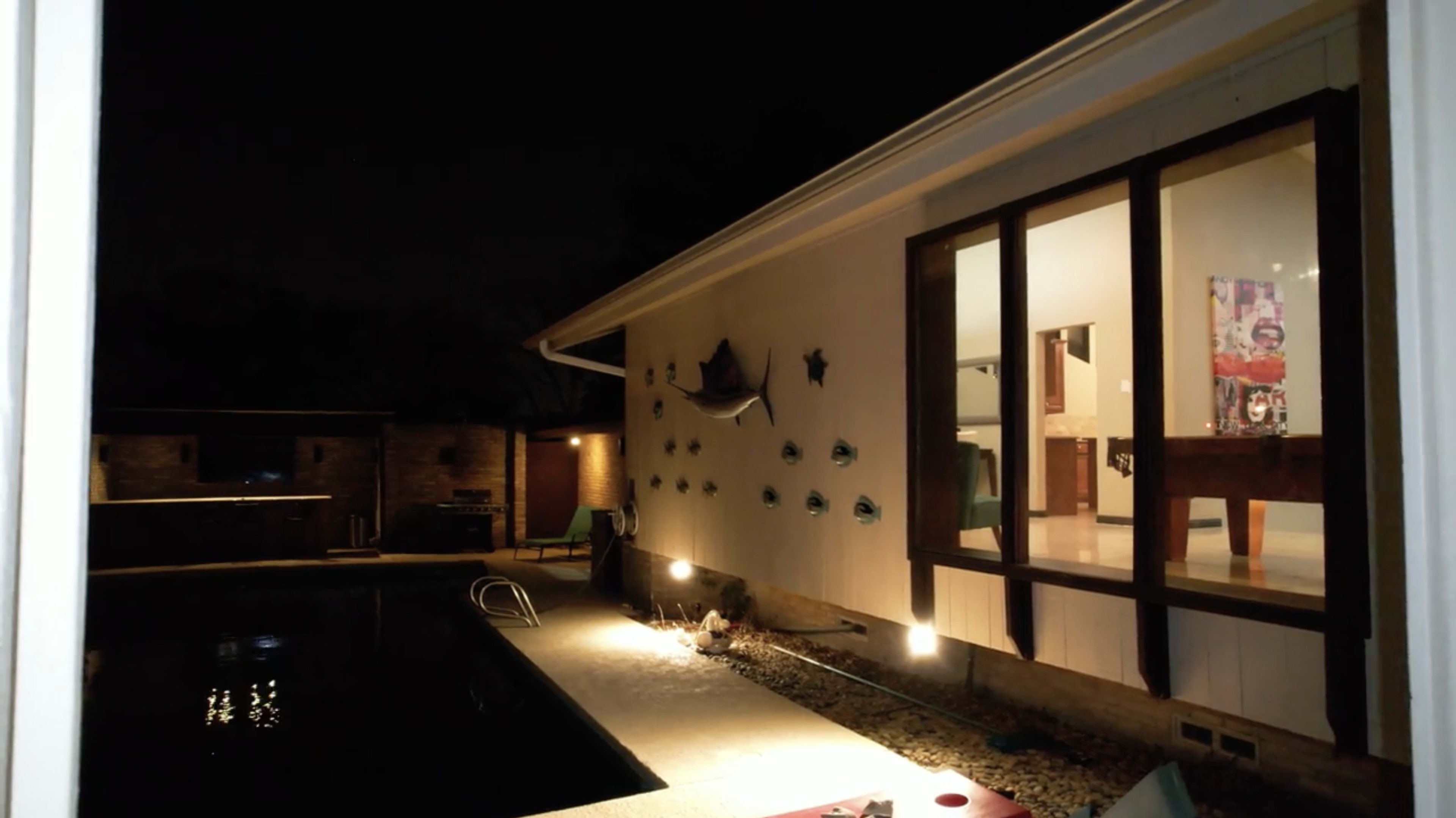 A well-lit patio area beside a swimming pool at night, with a view of a house featuring large windows and decorations on the wall.