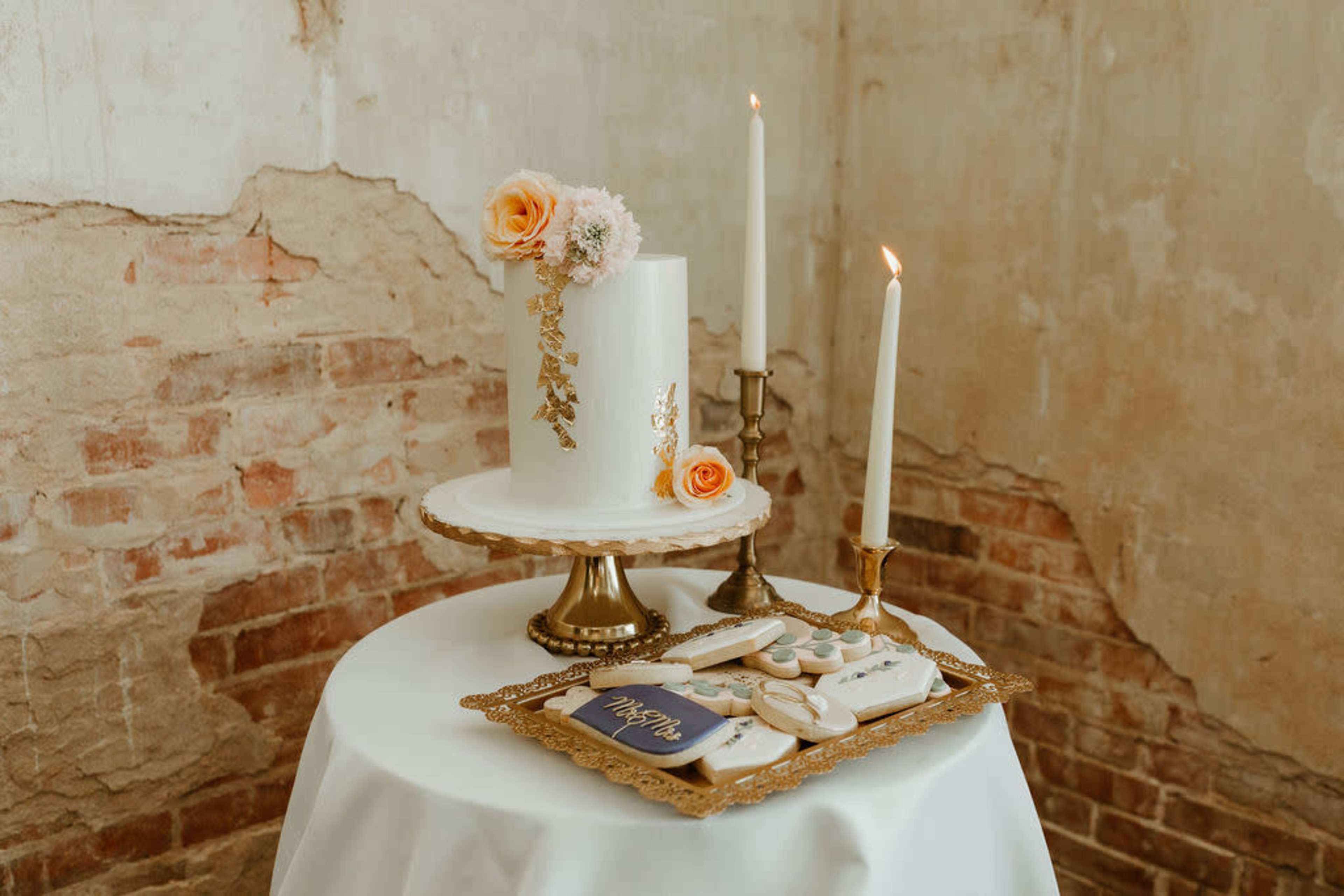 A white tiered cake is arranged on a pedestal with decorative flowers beside two lit candles and a tray of cookies on a table draped with a cloth.