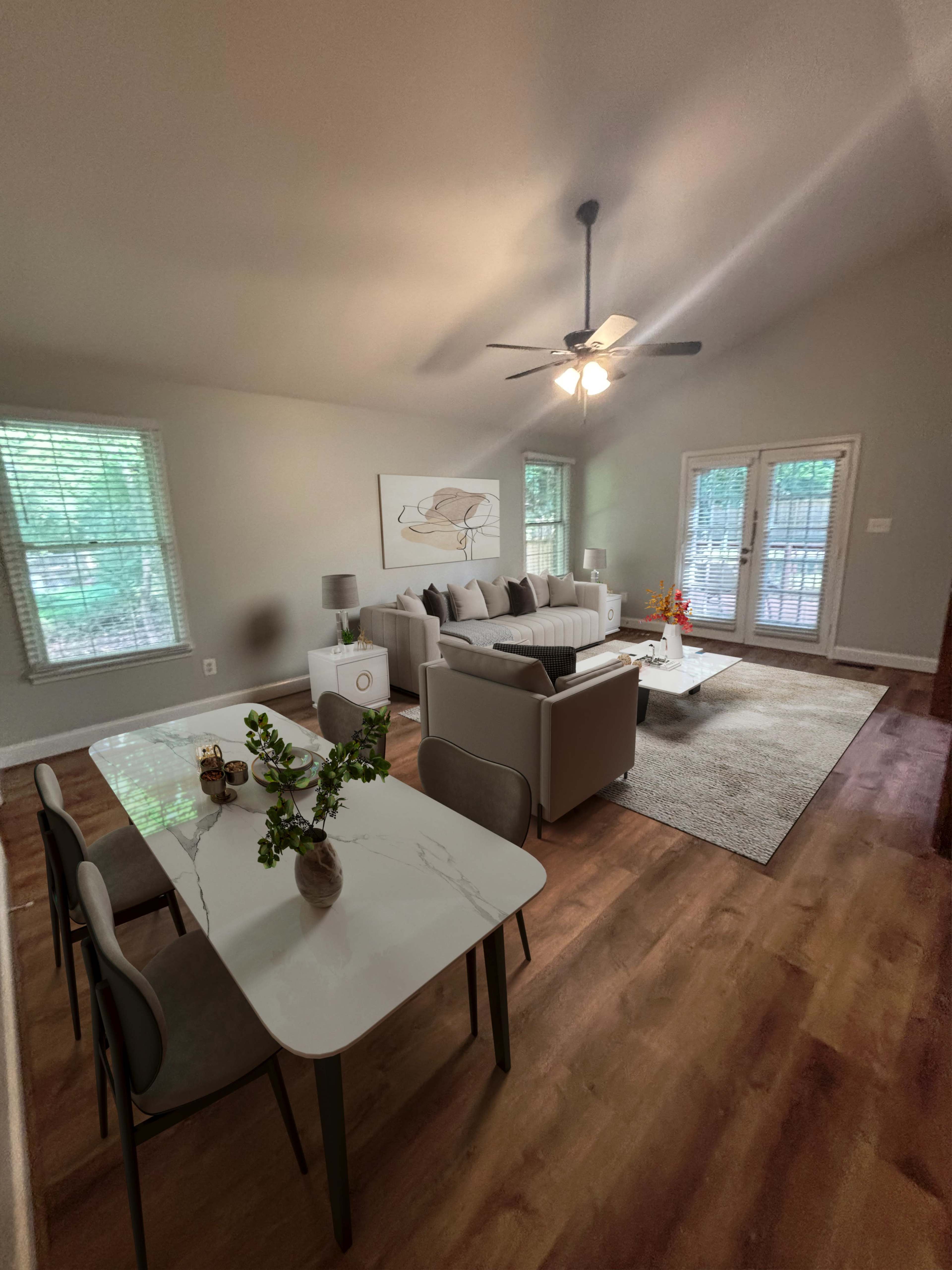 The image shows a bright, modern living room with a seating area, a dining table, and large windows allowing natural light to enter.