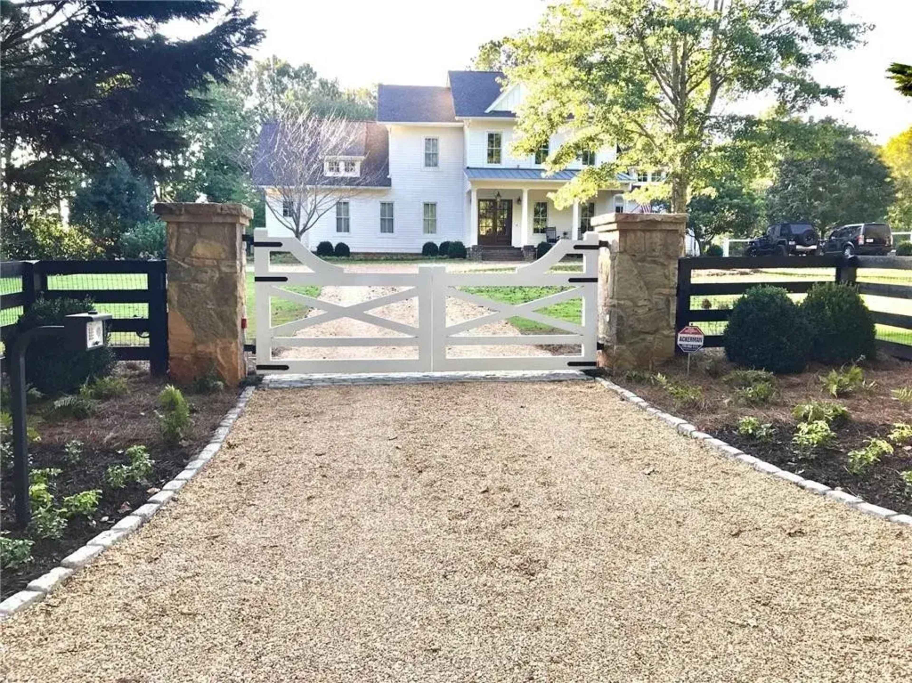 A white gate with stone pillars stands at the entrance of a gravel driveway leading to a large white house surrounded by trees and landscaping.