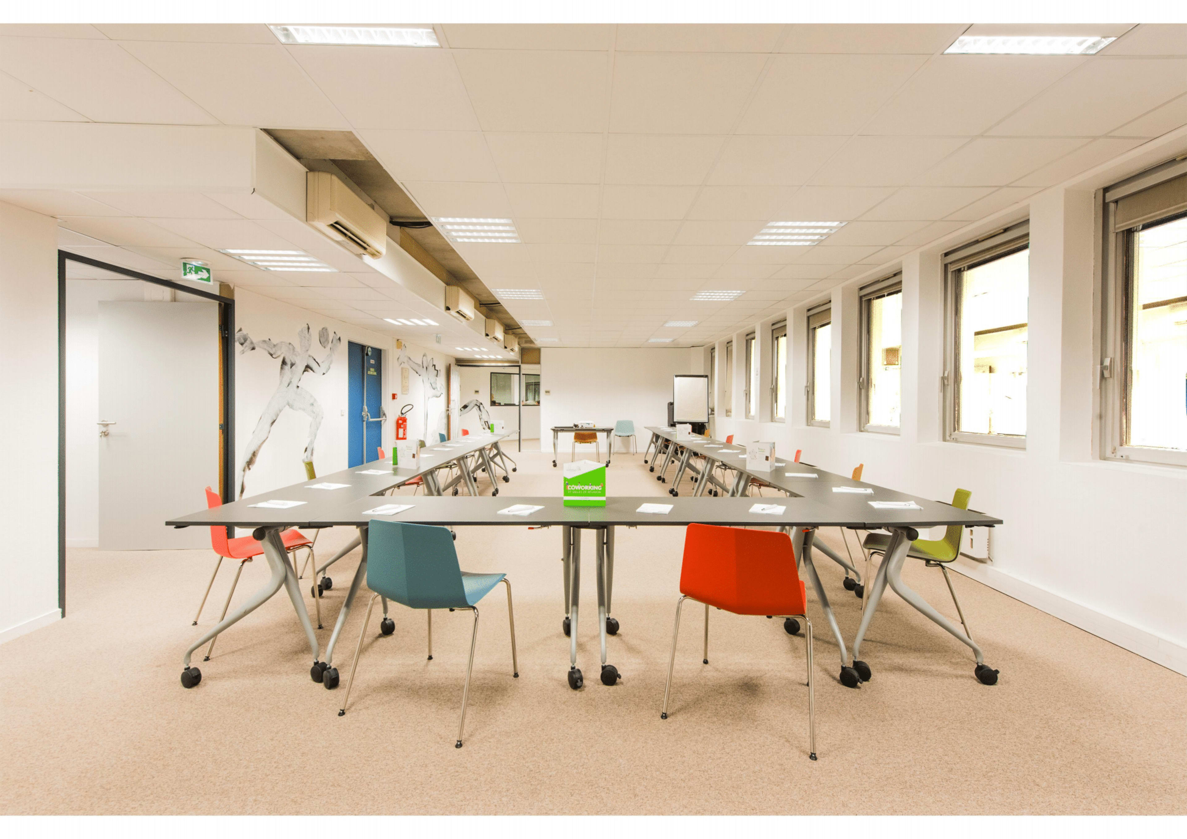 A modern meeting room features a circular arrangement of tables and colorful chairs with large windows and a whiteboard at the front.